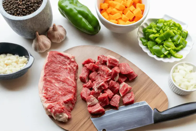 Raw steak and diced beef on wooden board surrounded by chopped vegetables and garlic cloves