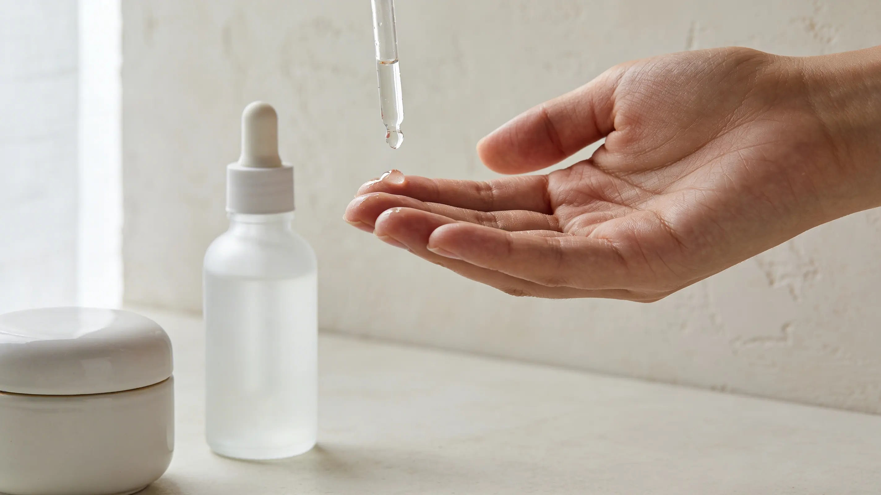 delicate, well-moisturized hand interacting with a few unbranded, minimalist skincare containers such as a frosted glass serum dropper and a smooth ceramic cream jar