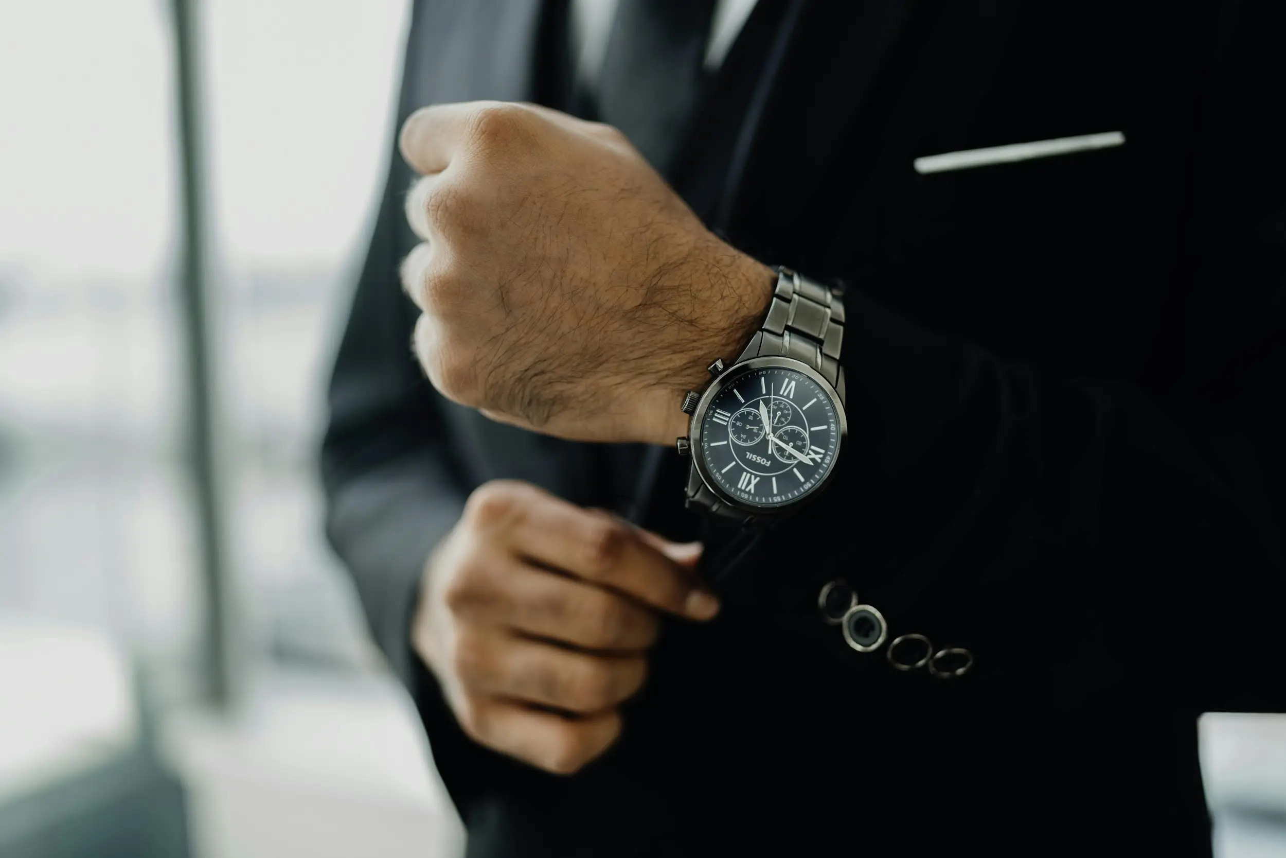 Man adjusting black wristwatch on left wrist, wearing a black suit indoors