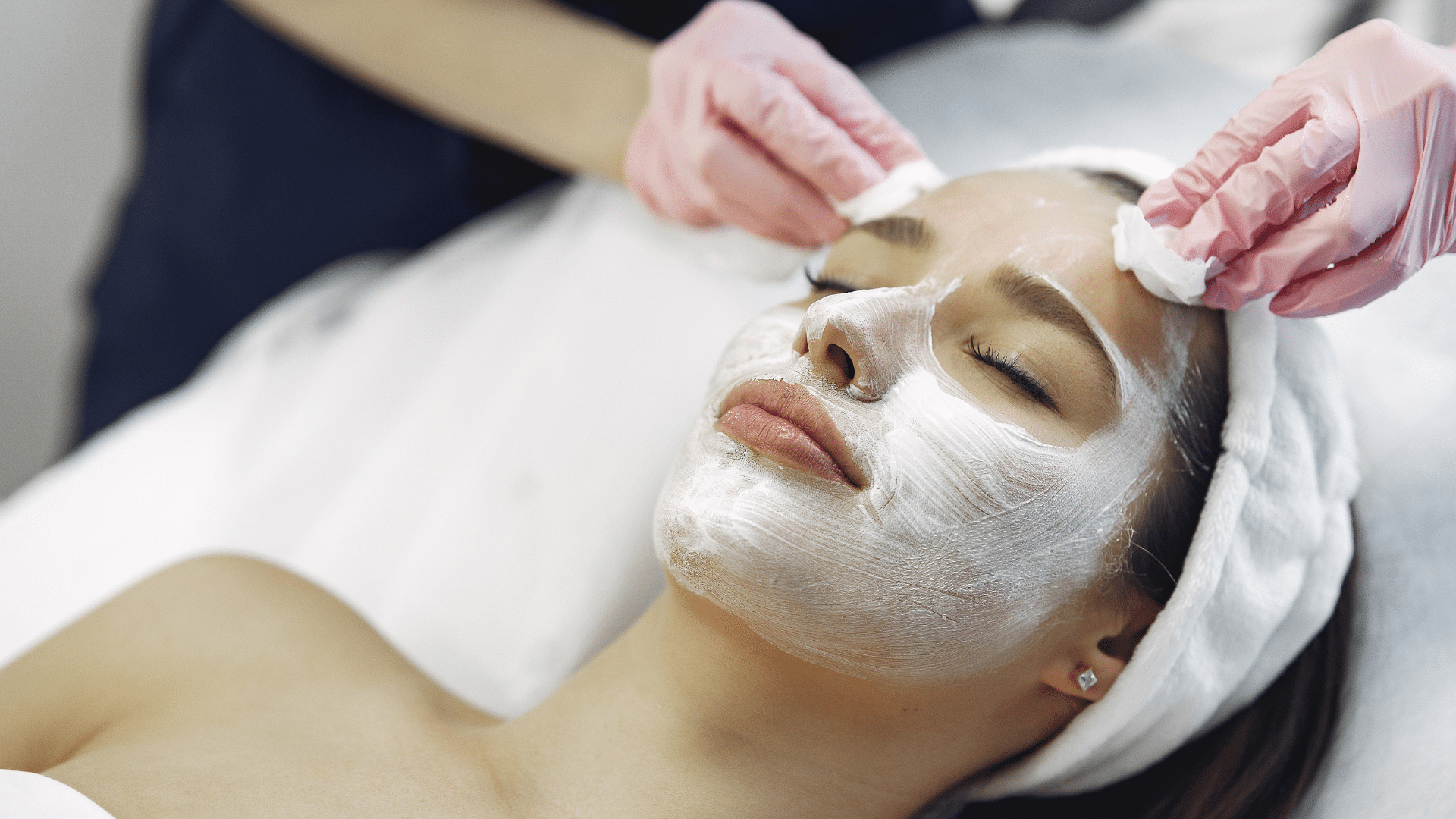 receiving facial treatment with mask applied while esthetician gently cleans skin using cotton pads in spa setting calm relaxing