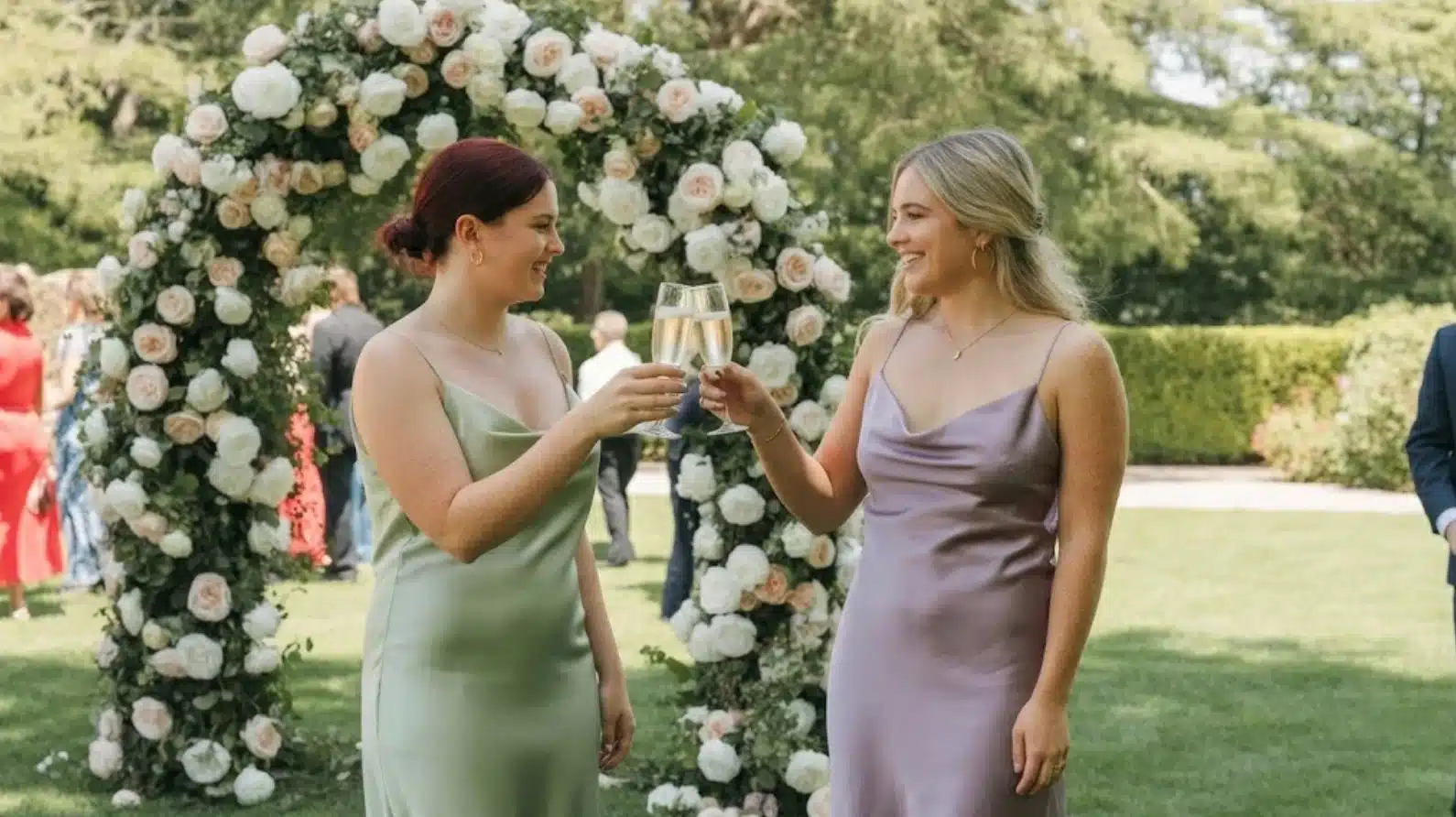 two women in satin dresses clinking champagne glasses beneath floral arch at outdoor garden wedding celebration