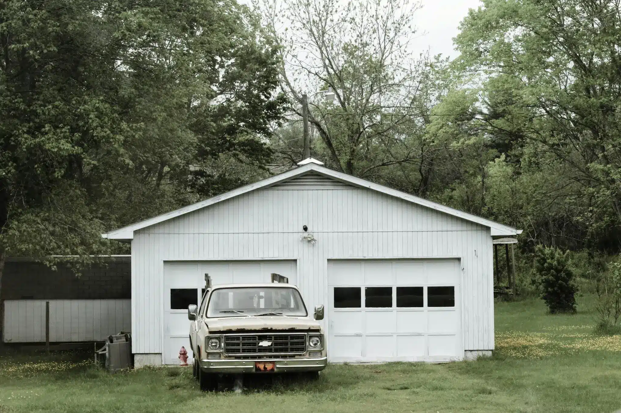 Old truck parked in front of white garage surrounded by trees and grass