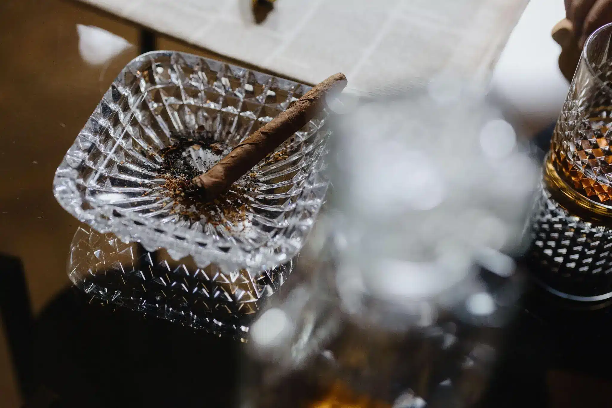 Cigar resting in a glass ashtray on reflective table with whiskey glass nearby