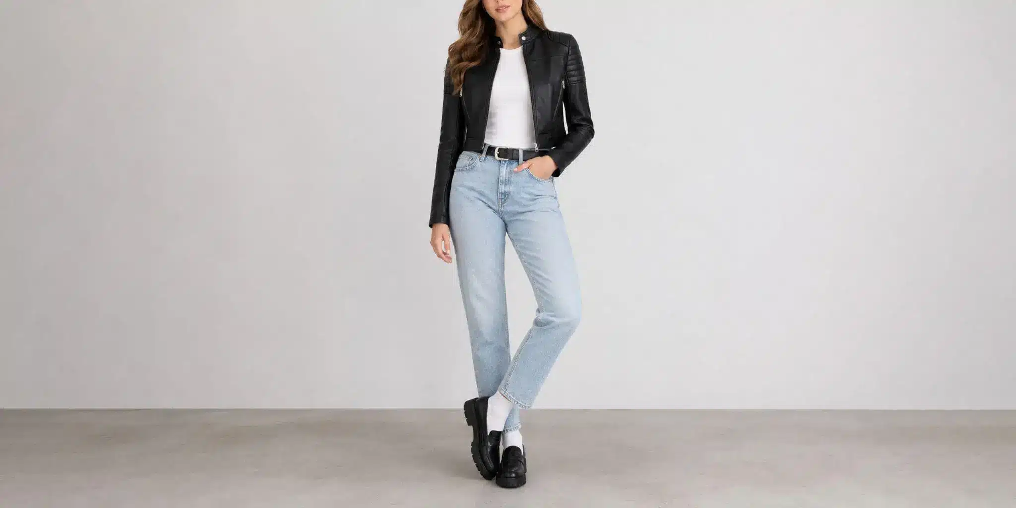 Woman wearing black leather jacket, white top, light blue jeans, black shoes in a minimal studio setting