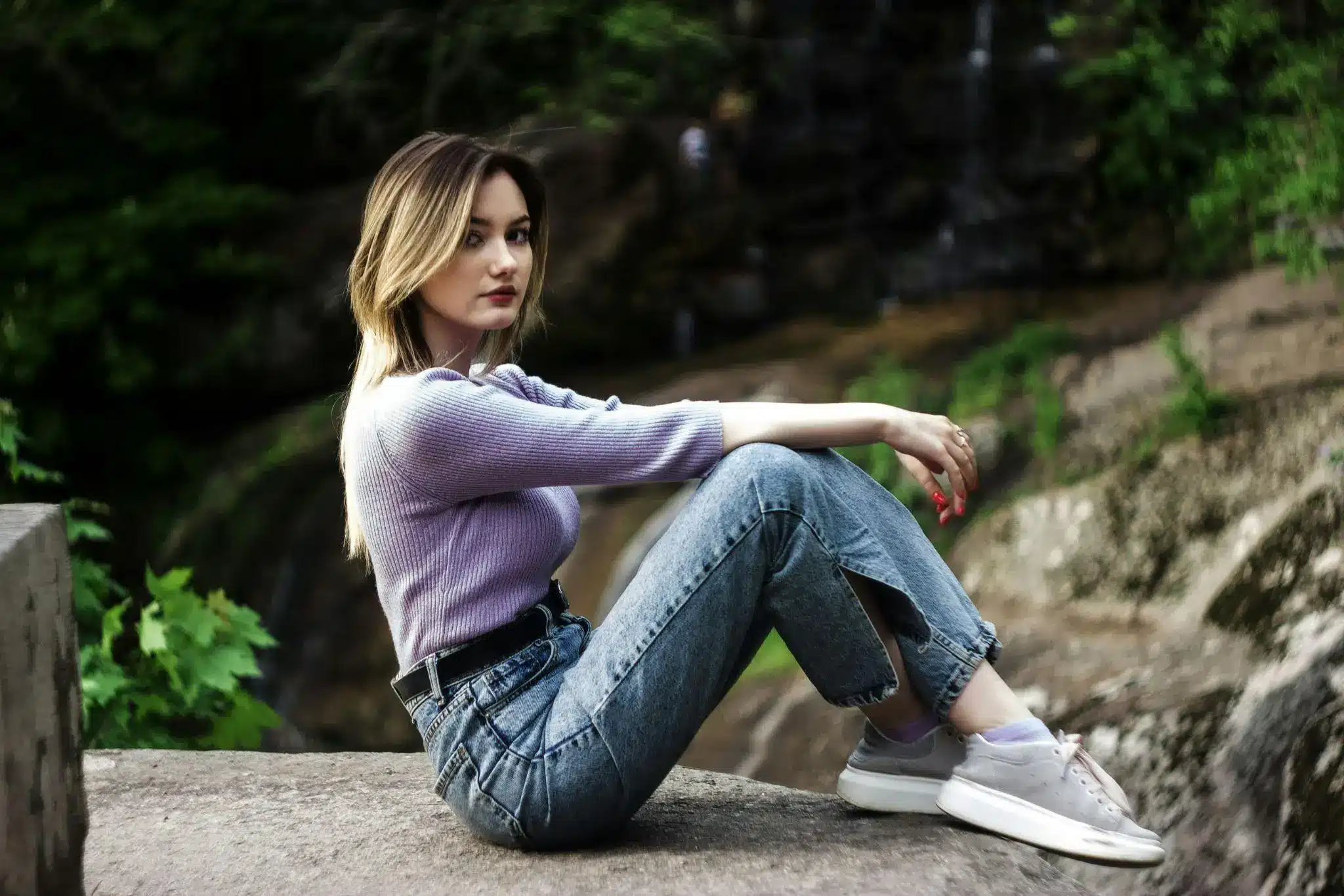 Woman in purple sweater and jeans sitting on rock in wooded area
