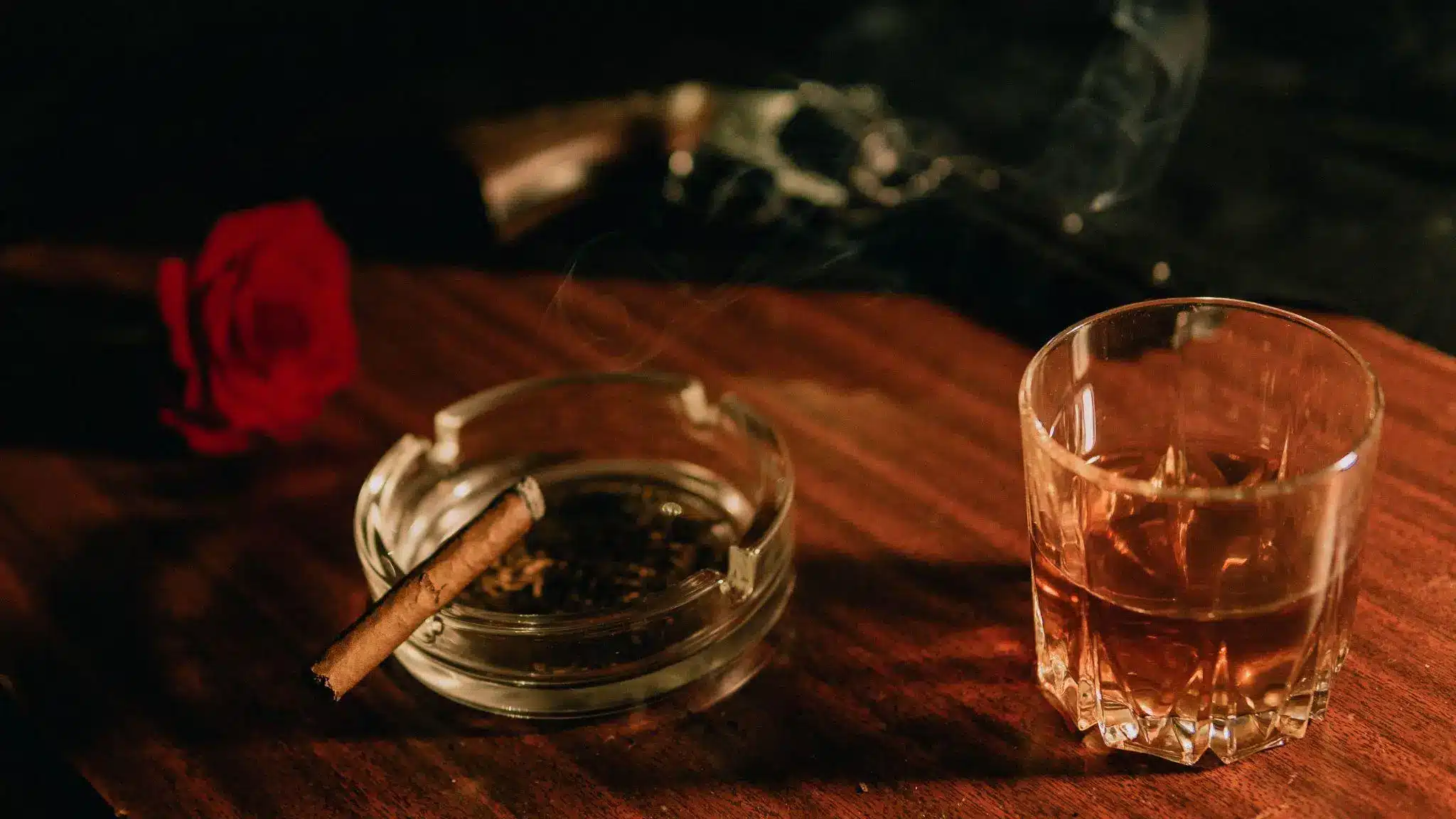 Cigar and whiskey glass on wooden table with red rose nearby in dim lighting