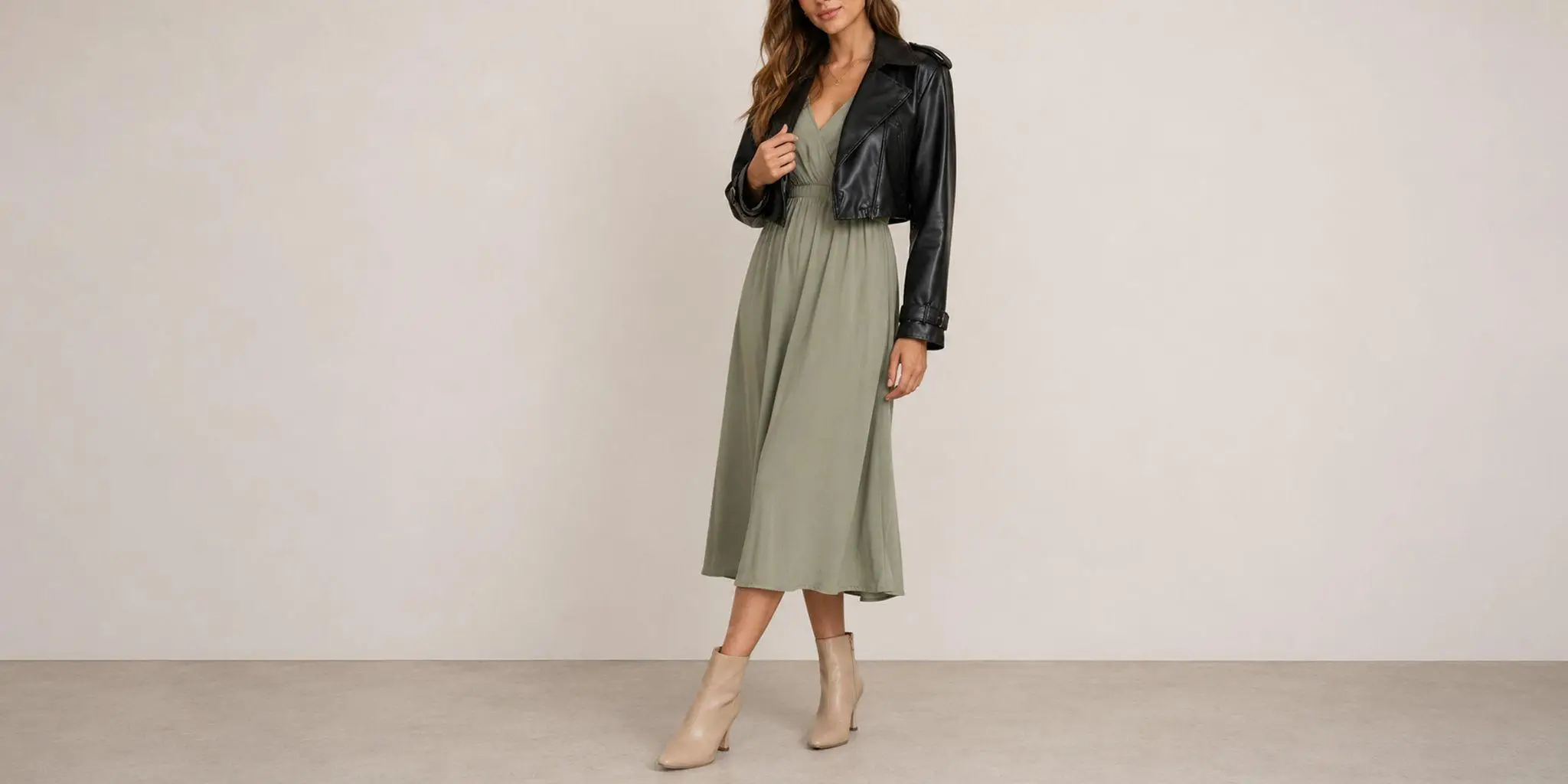 Woman in olive green dress and black leather jacket standing indoors against beige backdrop