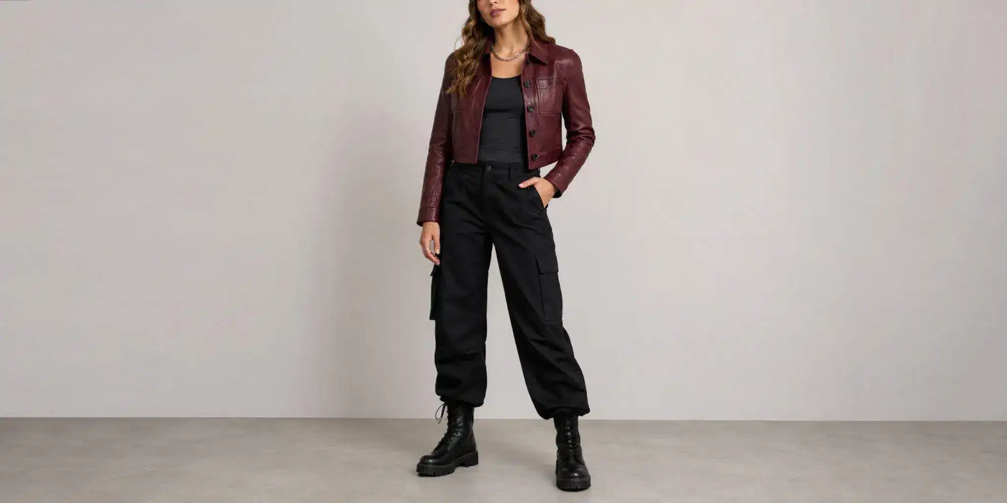 Woman in burgundy leather jacket and black cargo pants standing against gray wall