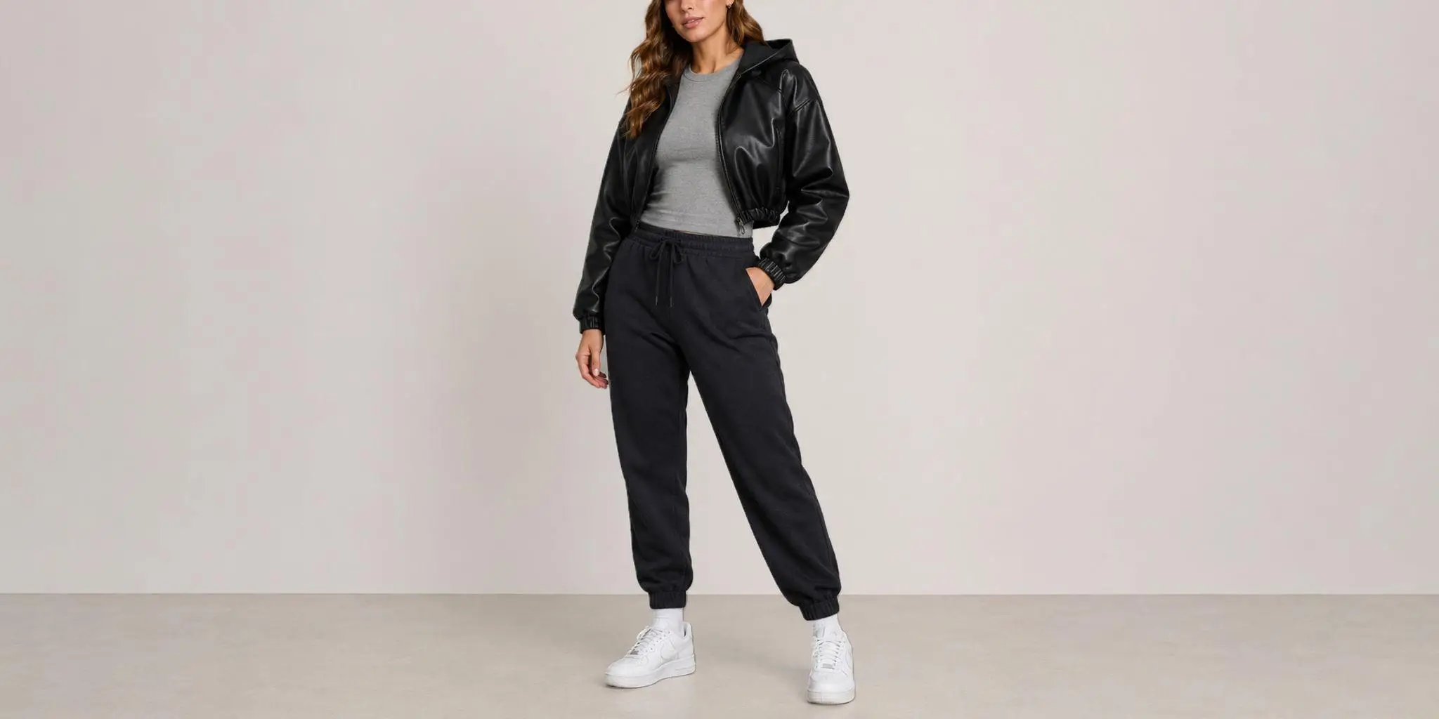 Person wearing gray top, black leather jacket, dark joggers, and white sneakers against plain background