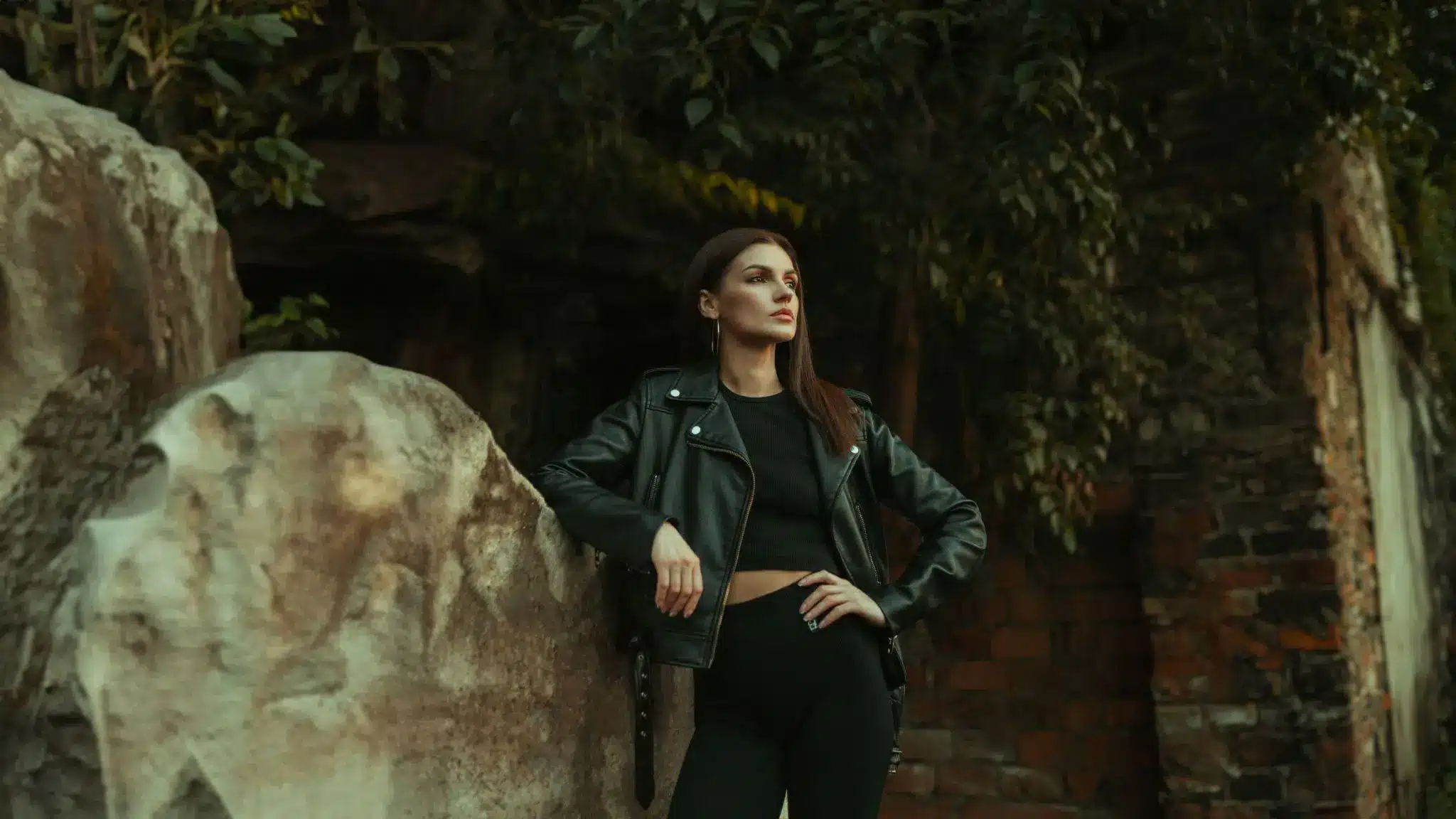 Woman in black leather jacket leaning against a stone wall in an outdoor setting