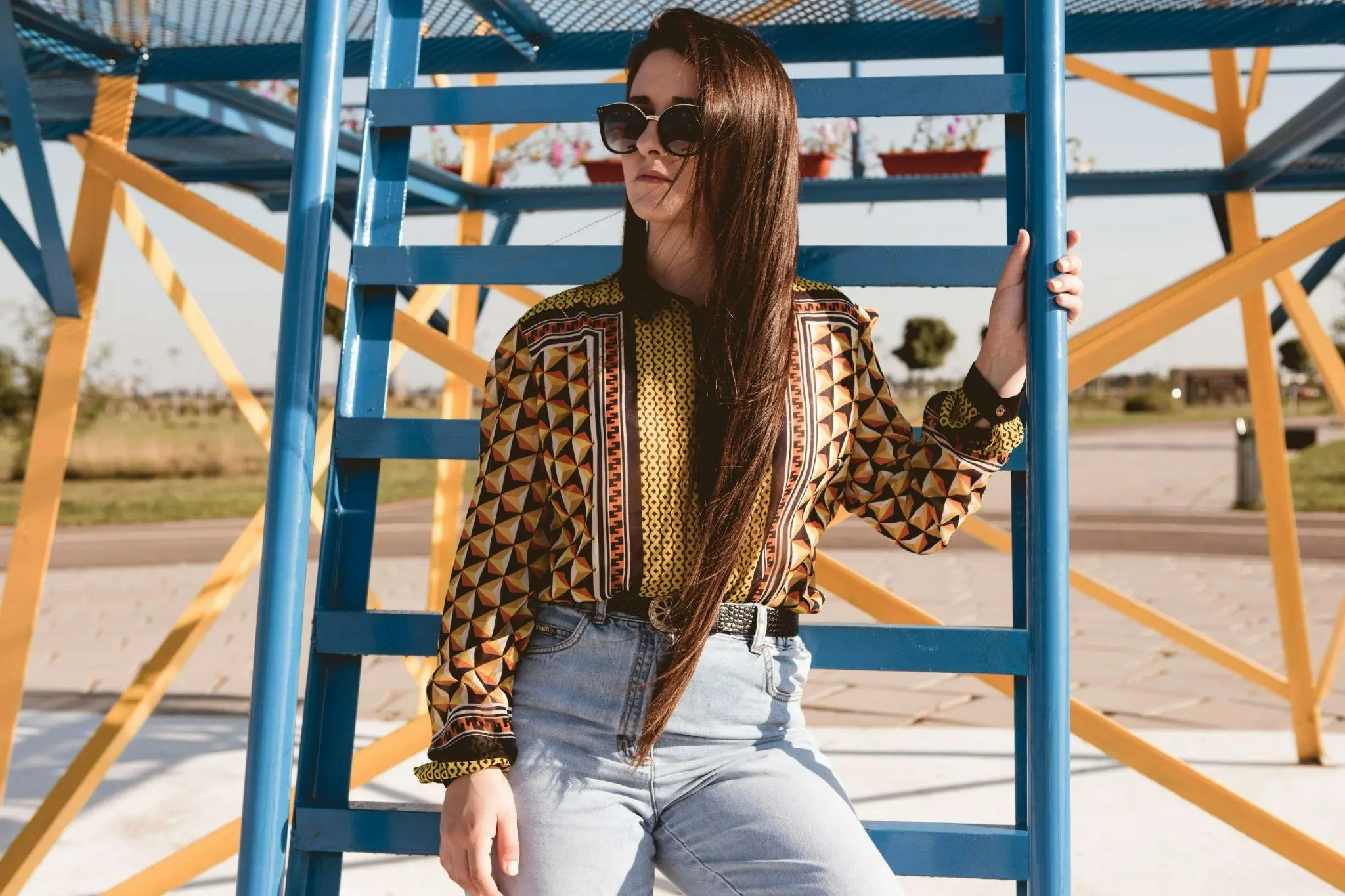 Woman in geometric patterned shirt and sunglasses standing by blue and yellow metal structure outdoors