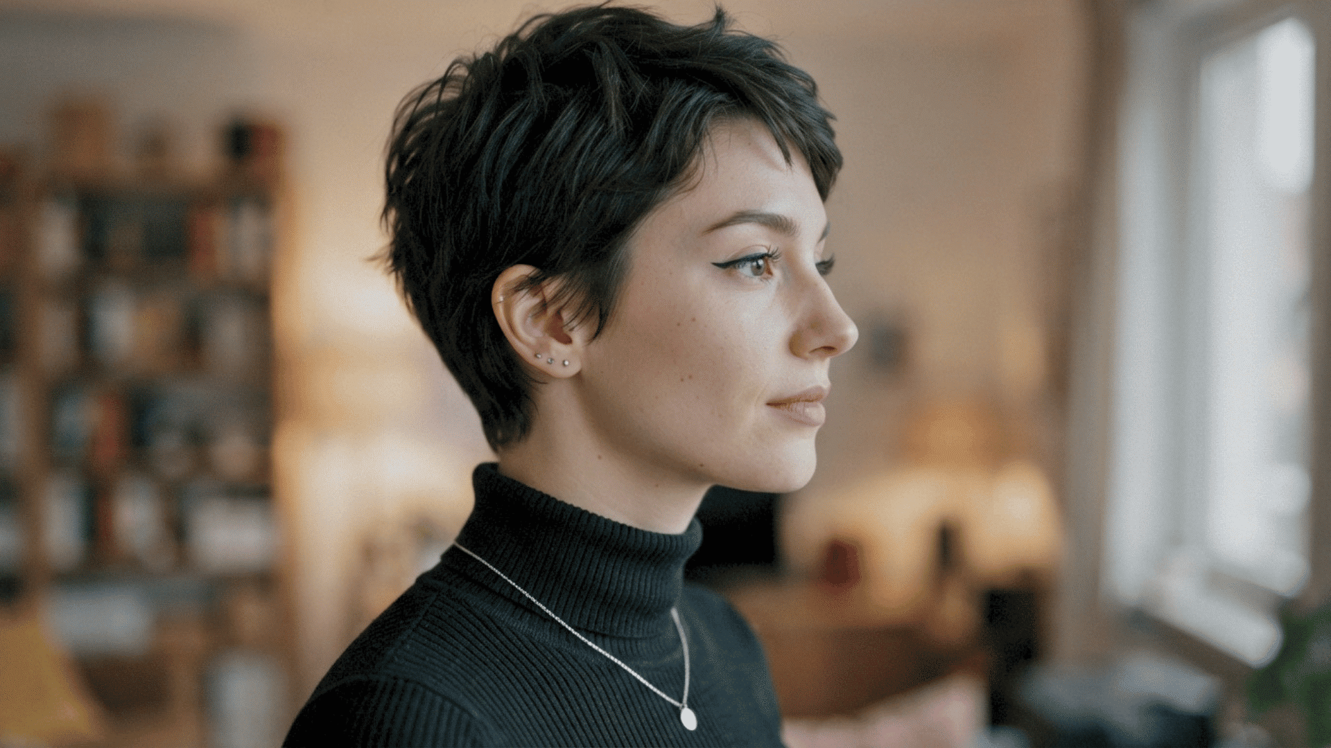Person with short pixie haircut wearing a black turtleneck and necklace in a side profile portrait