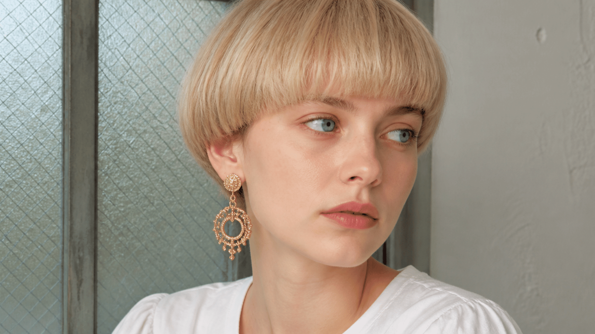 Person with short blonde bowl haircut and statement earrings posing in a close-up portrait