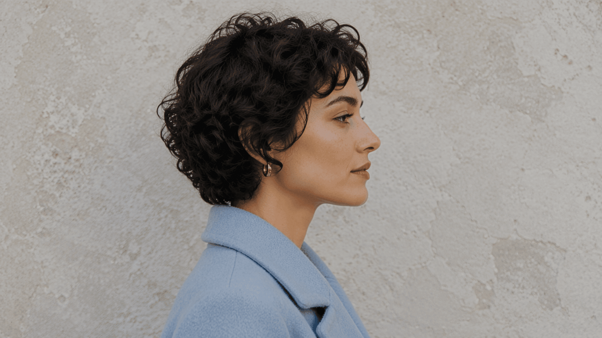 Person with curly pixie cut haircut posing in side profile against a textured wall
