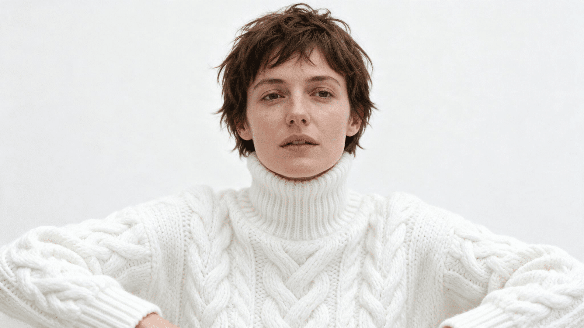 Person with long pixie haircut wearing a white cable knit sweater in a minimalist portrait