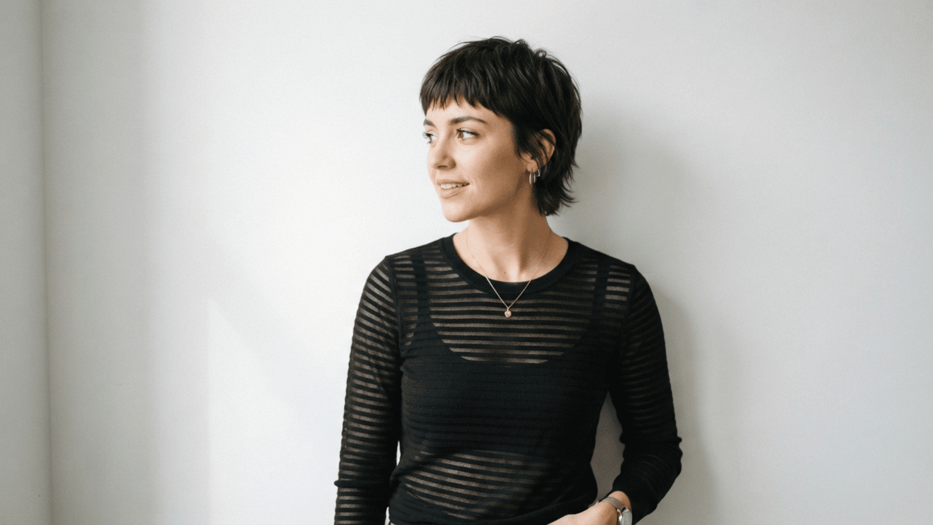 Person with short haircut and micro bangs wearing a black striped top in a minimalist portrait