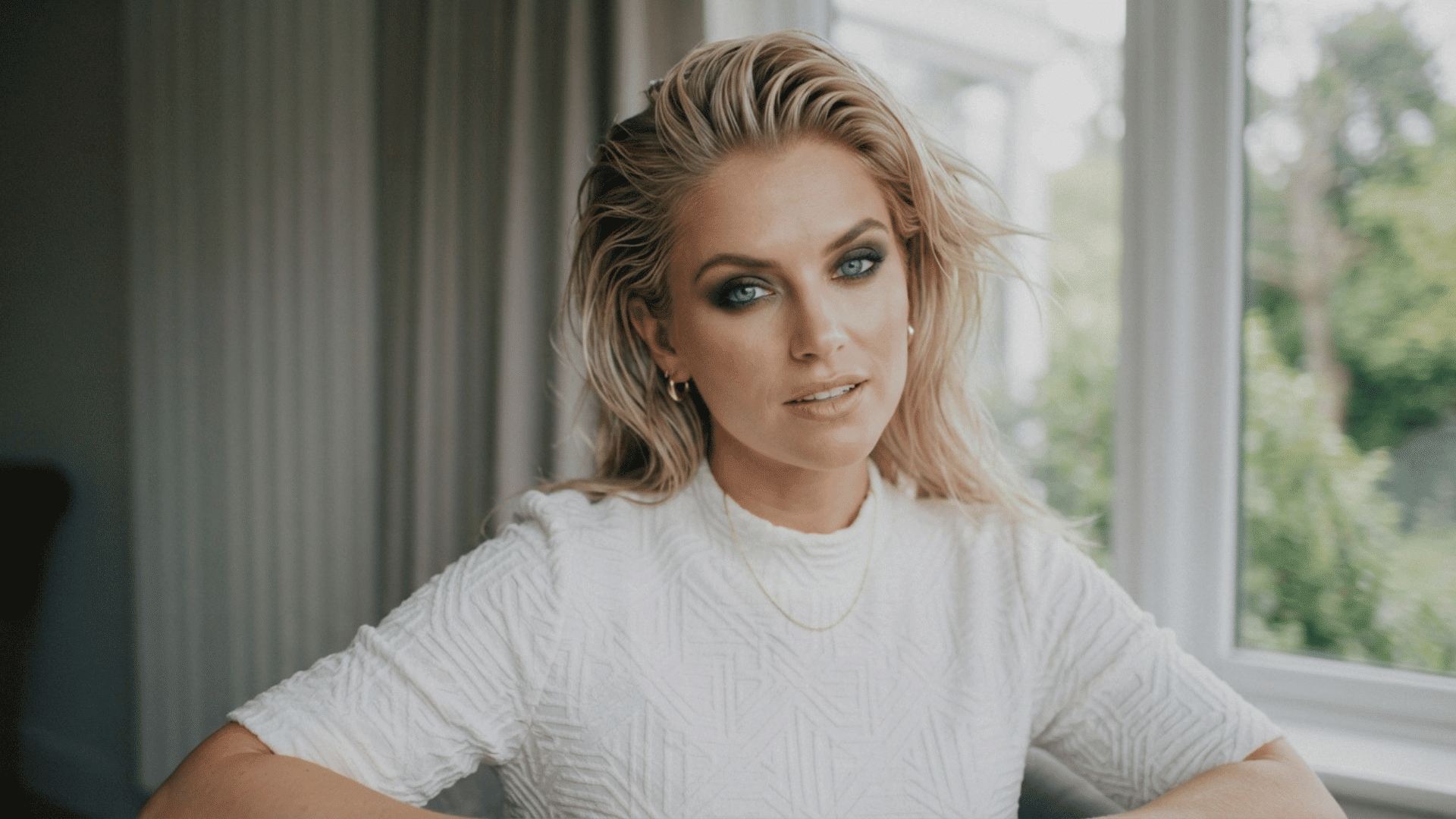 Person with slicked-back blonde hair wearing a white textured sweater in an indoor portrait