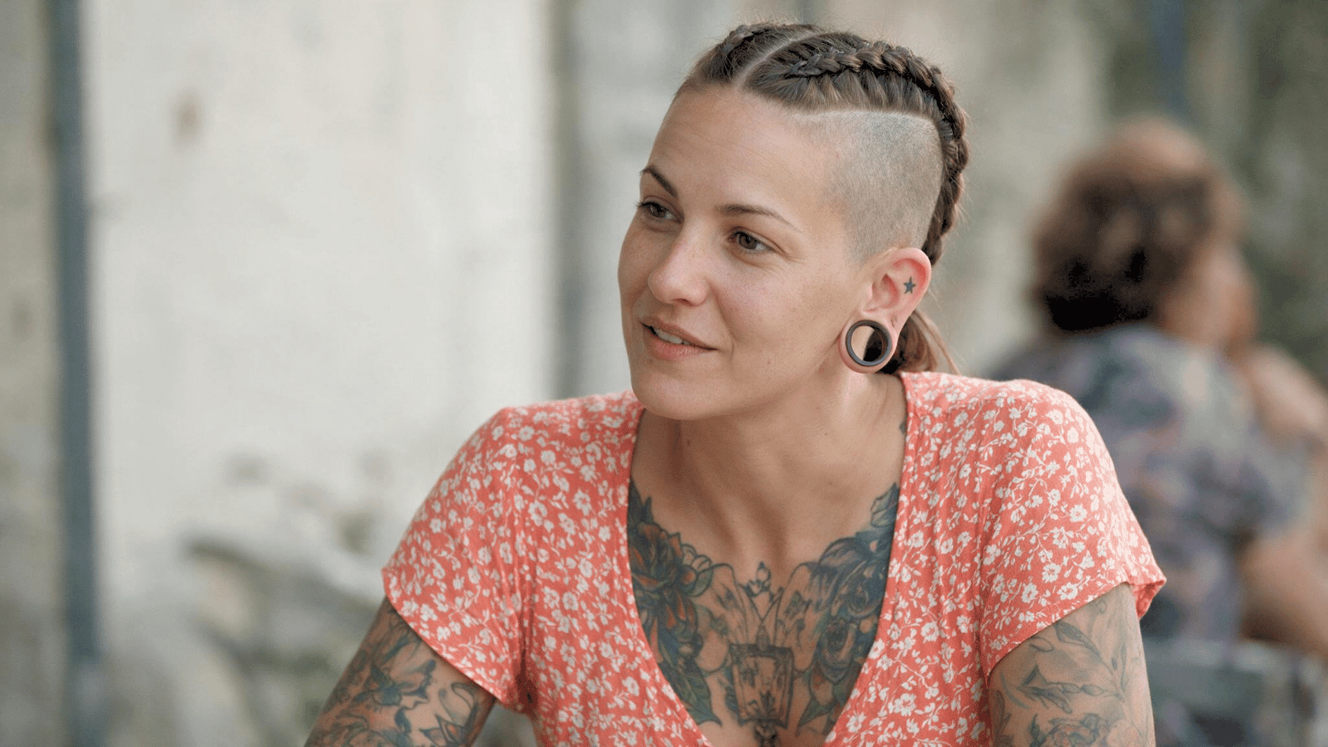 Person with braided undercut hairstyle, tattoos, and earrings posing outdoors in a floral top