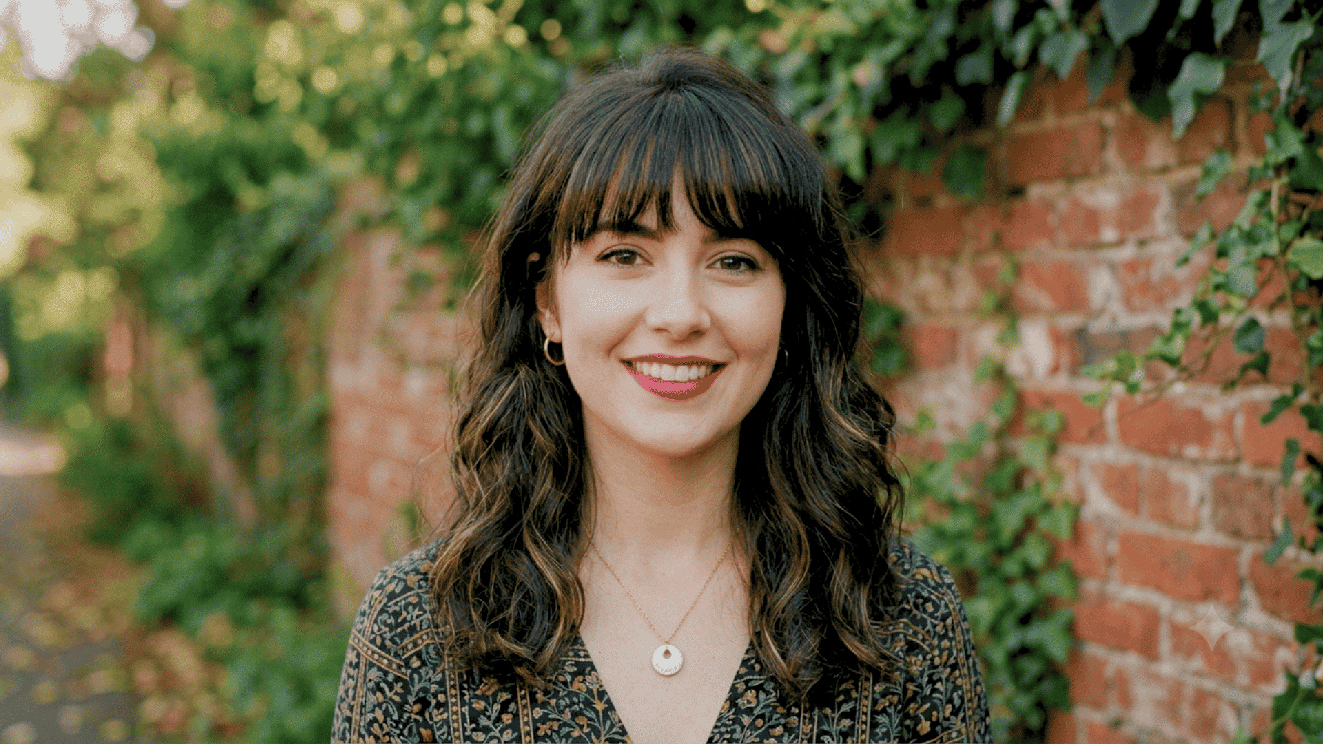 Person with a wolf cut haircut and bangs smiling outdoors beside a brick wall and greenery