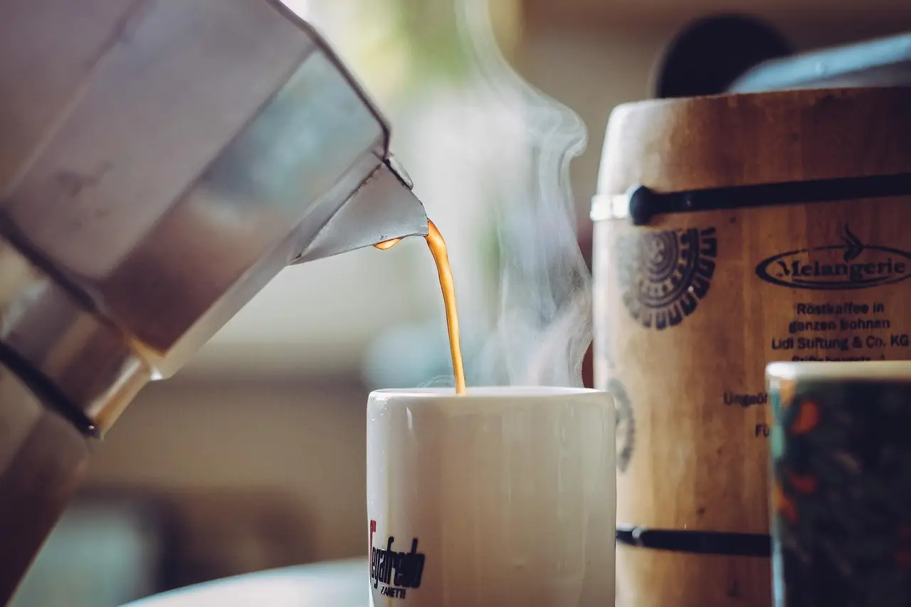 Steaming coffee being poured from moka pot into white mug in cozy kitchen setting