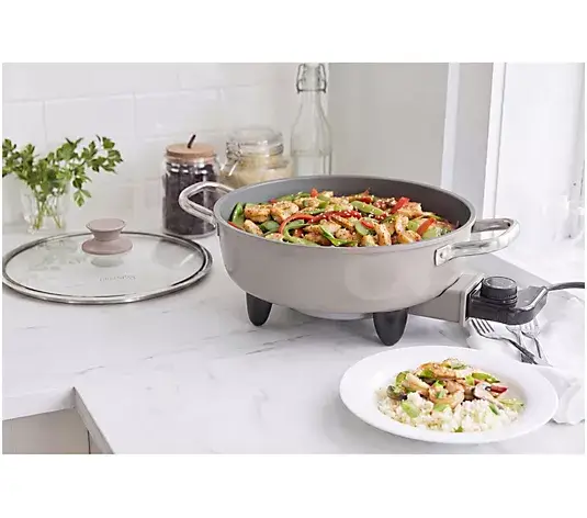 Electric skillet cooking stir fry on white kitchen countertop with plate of rice and vegetables nearby