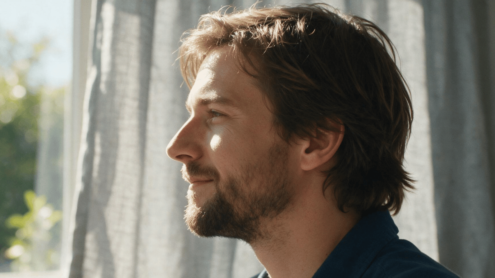 Man with medium-length hair sitting near a window in natural sunlight.