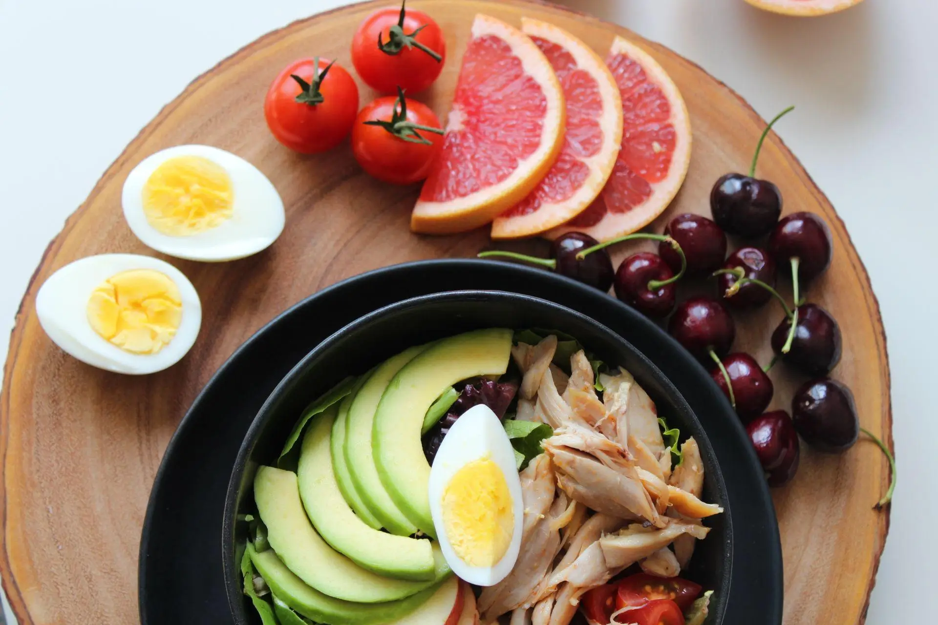 Salad bowl with avocado, chicken, and boiled egg on wooden platter surrounded by fruits