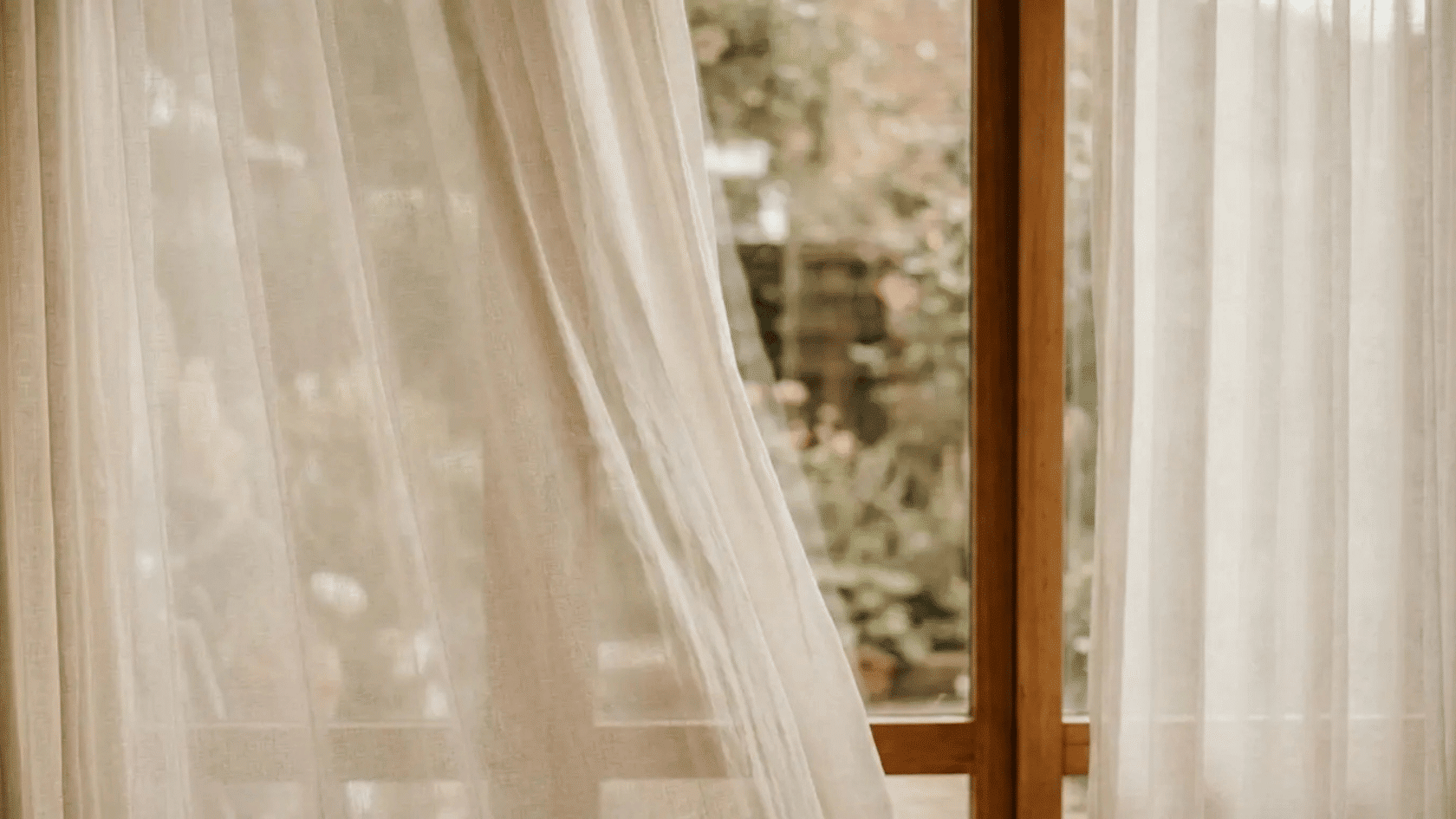 White sheer curtains softly filtering natural light beside a wooden window frame.