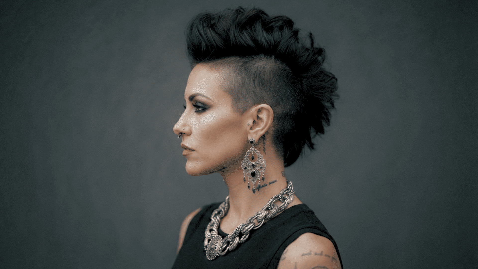 Person with mohawk hairstyle, earrings, and tattoos posing in a side profile portrait