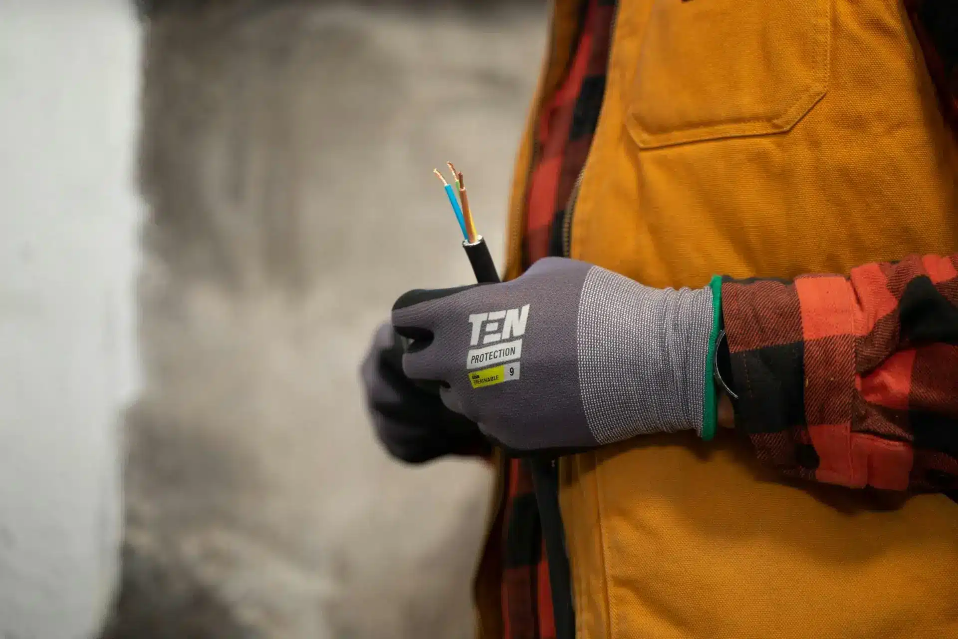 Person holding electrical wire with gloved hand in workshop wearing plaid shirt and yellow vest