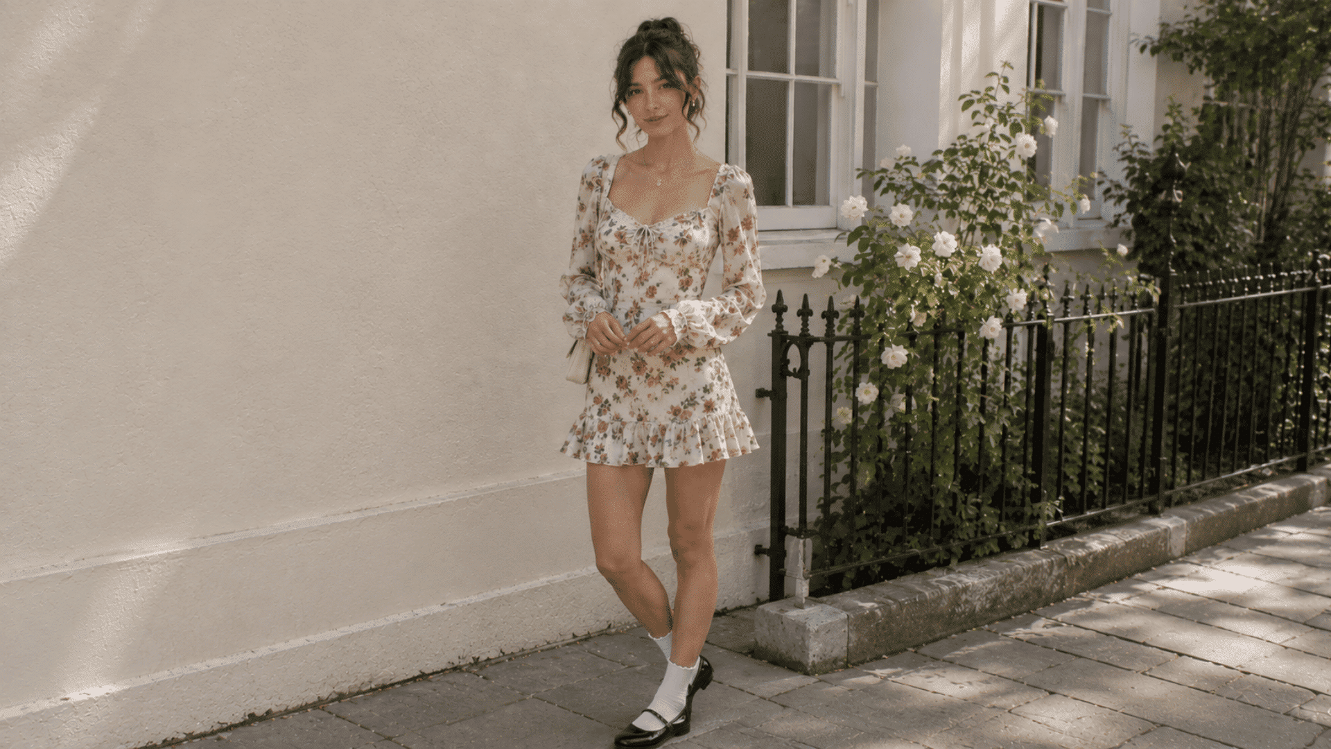 a woman wearing a floral mini dress with long sleeves and mary jane shoes standing beside a white rose garden