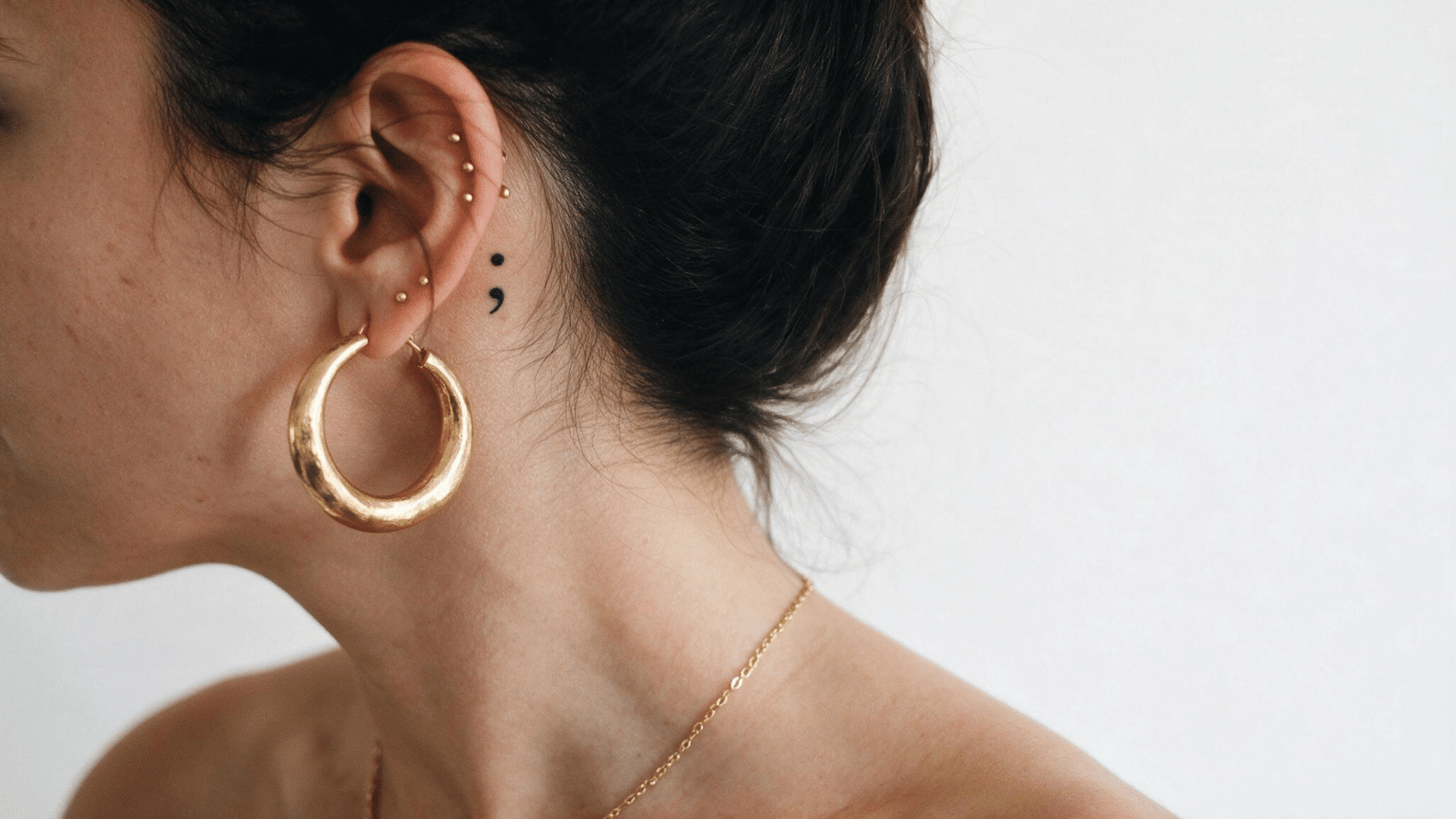 black semicolon tattoo behind a woman’s ear, paired with gold hoop earrings and multiple ear piercings
