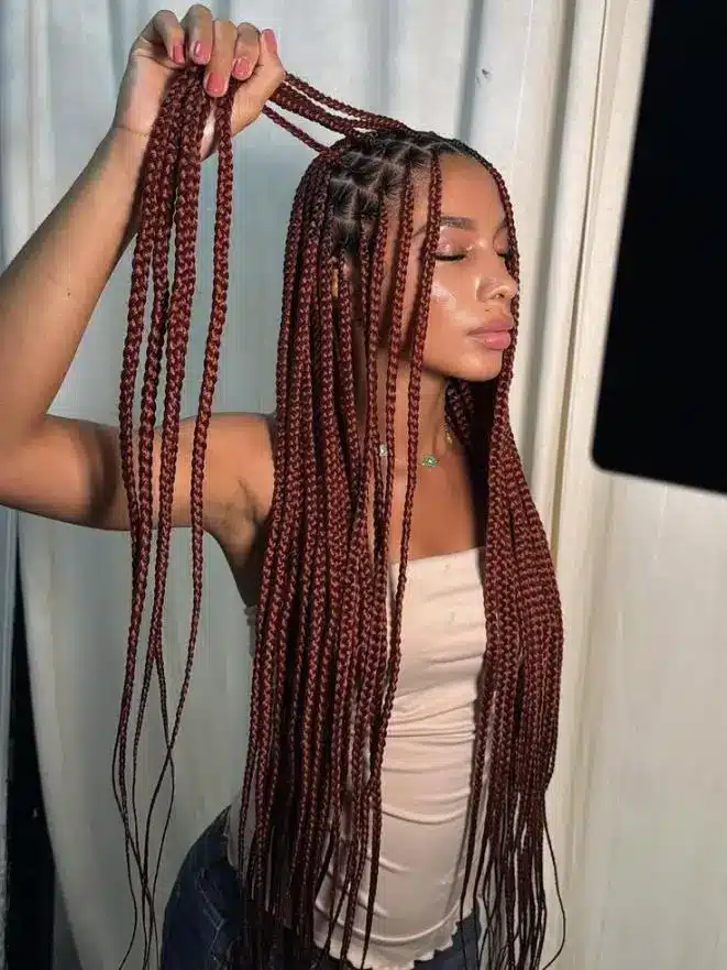 Person with long, detailed red braids in indoor setting with white curtain backdrop