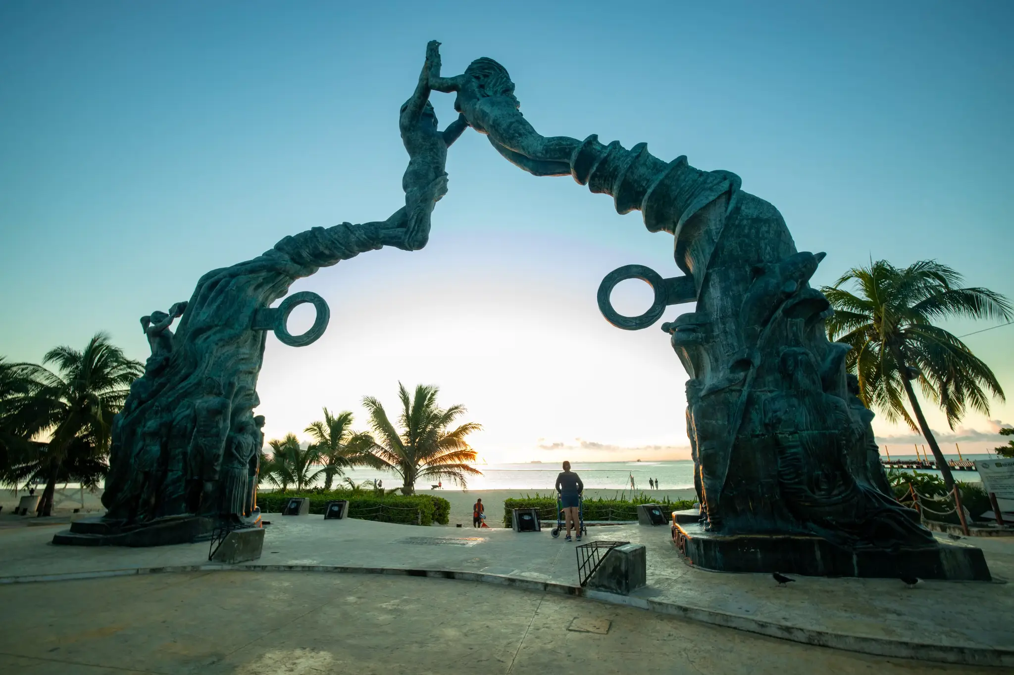 Sculptural arch with intertwined figures at sunset on a beach with palm trees