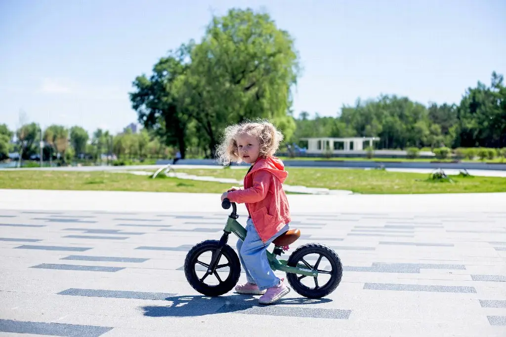 Child wearing red jacket riding balance bike on paved park path