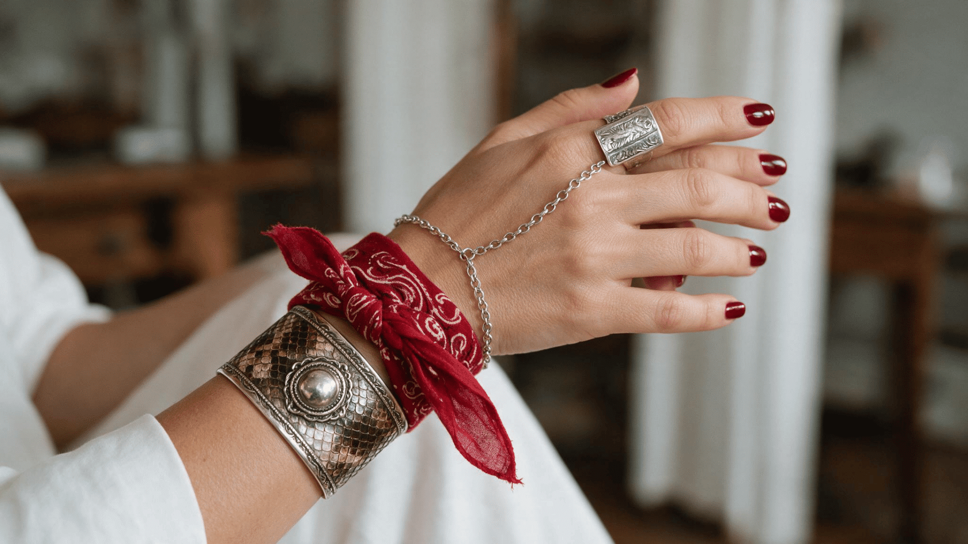 close-up of a red paisley bandana tied around a woman’s wrist, paired with silver bracelets, a hand chain, red nails, and an engraved ring.