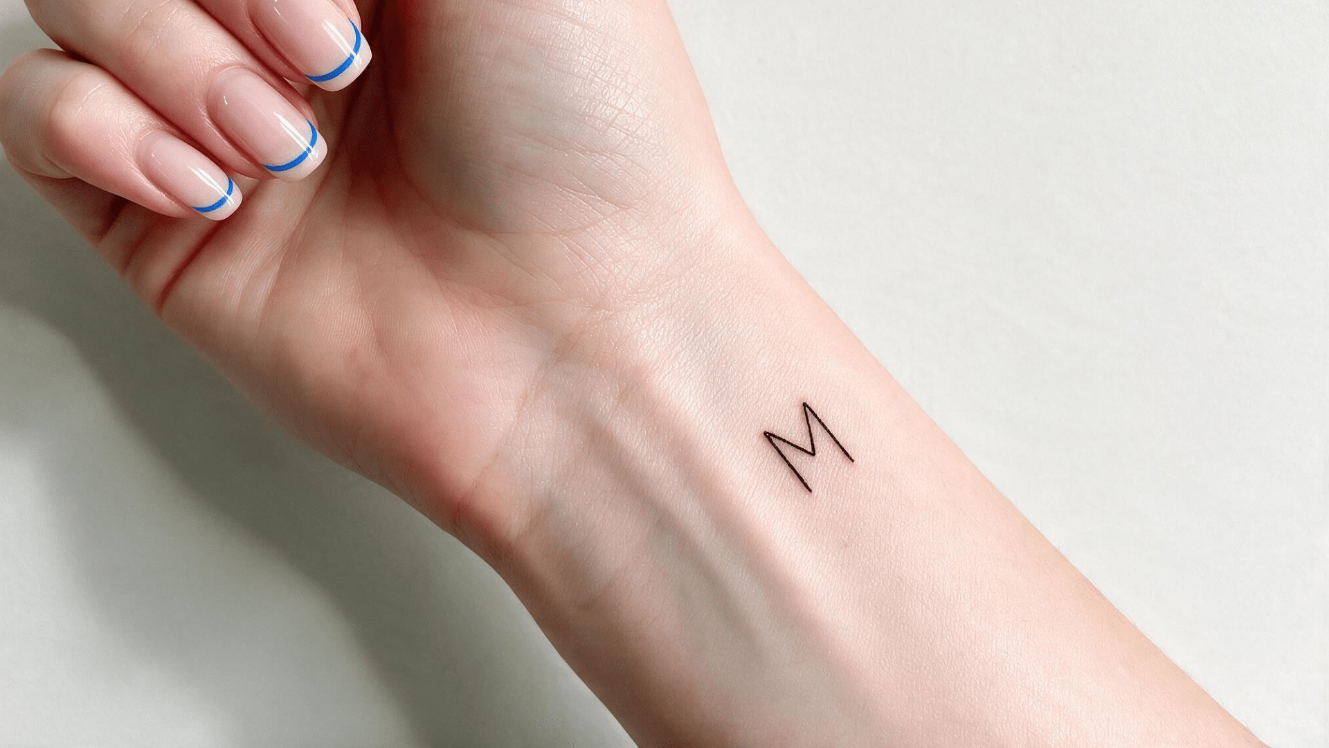close-up of a wrist with a small minimalist letter M tattoo and blue French tip nails.