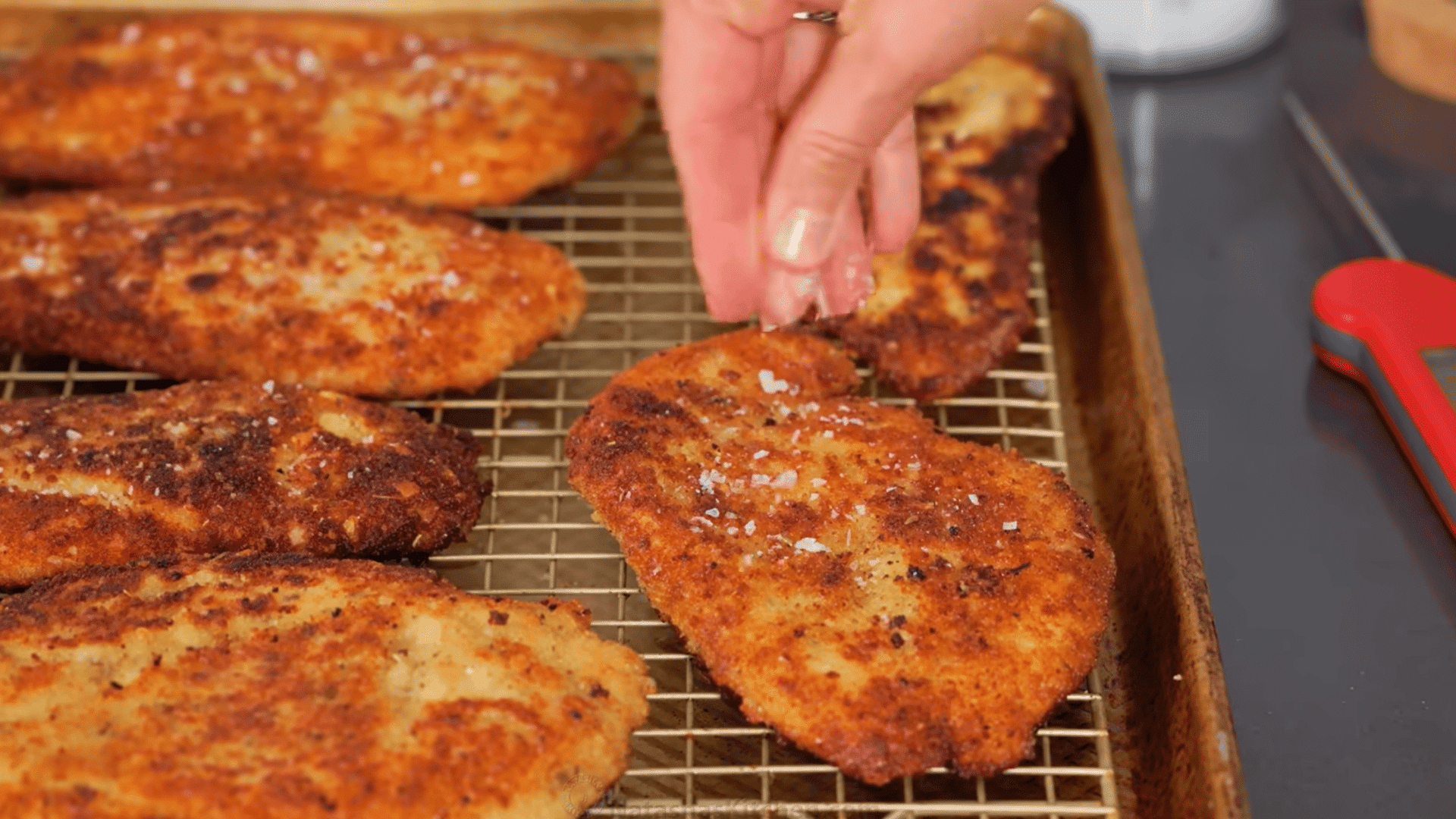 crispy chicken cutlets draining on a wire rack while being seasoned with flaky salt.