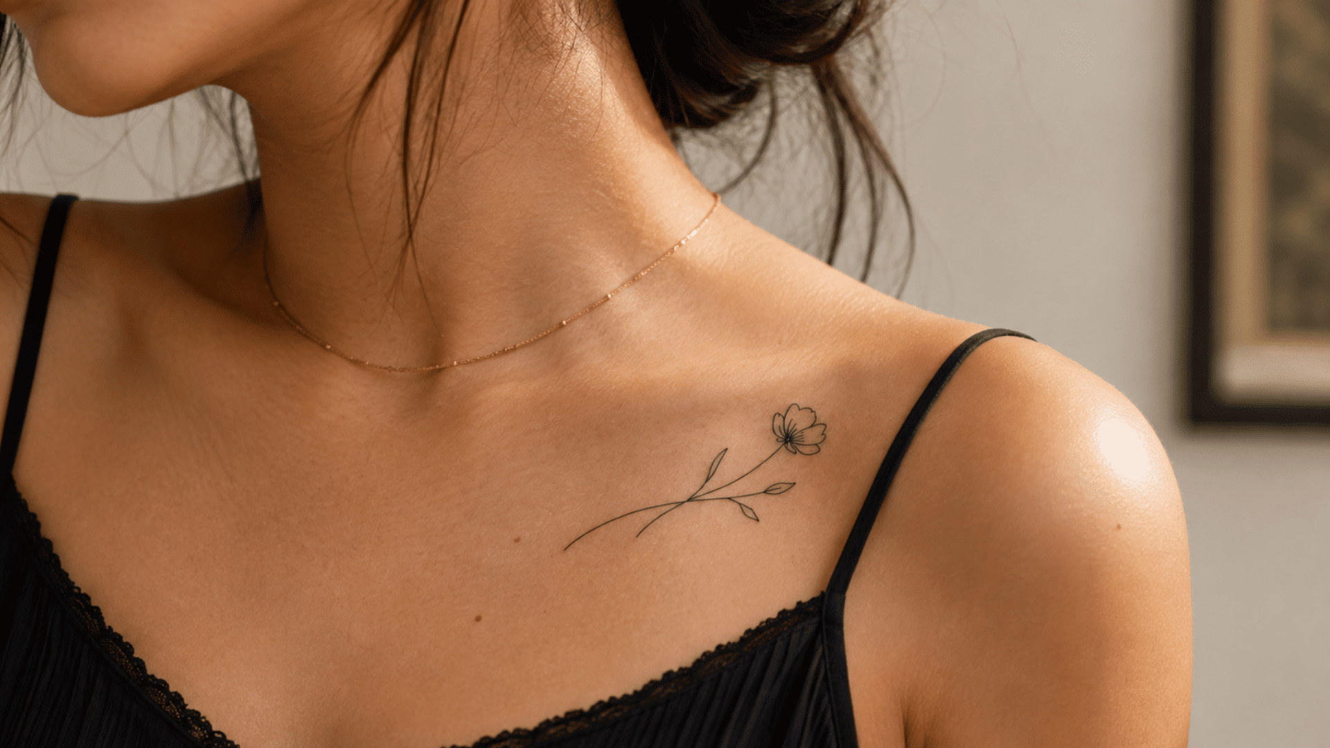 fine-line flower tattoo with a thin stem and leaves near the collarbone of a woman wearing a black camisole.