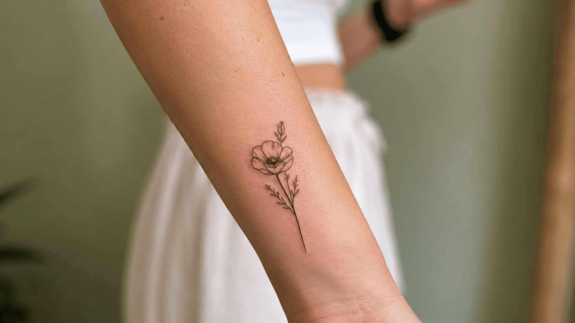 fine-line poppy flower tattoo with a stem and small leaves on the inner forearm.