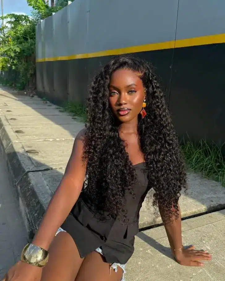 Woman with long curly hair sitting on sidewalk in casual outfit under bright sunlight