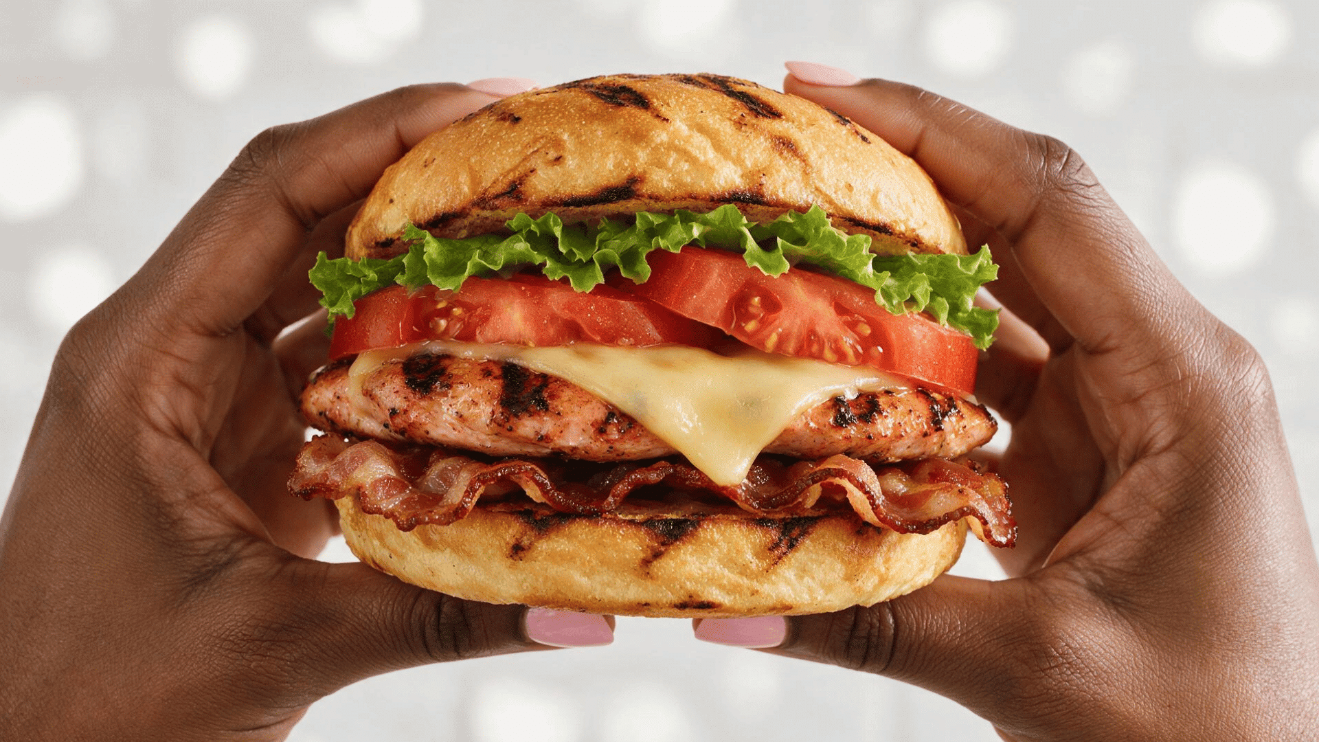 grilled chicken sandwich with lettuce, tomato, cheese, and bacon held in both hands.