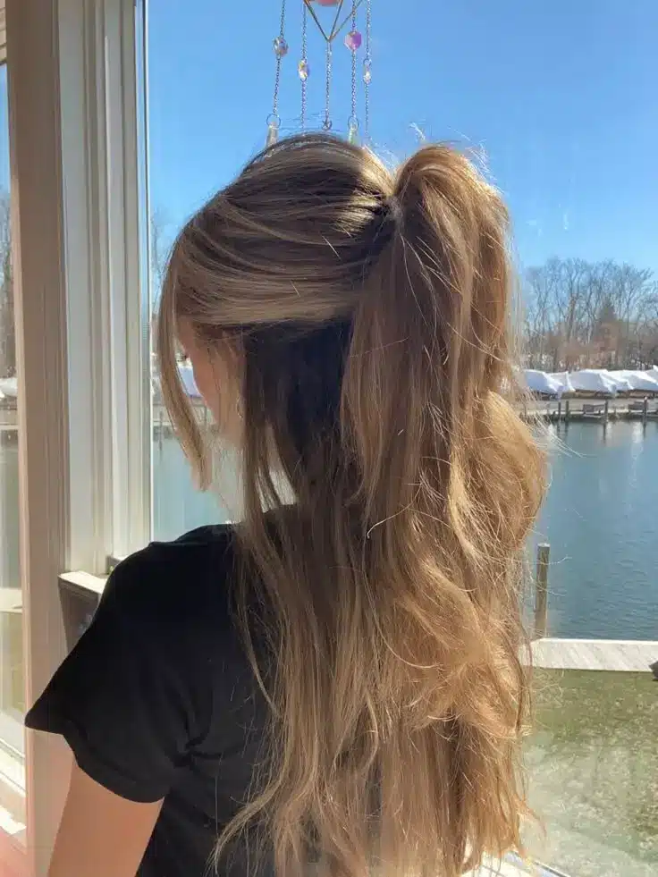 Woman with long wavy blonde hair in ponytail facing window overlooking sunny waterfront scene.