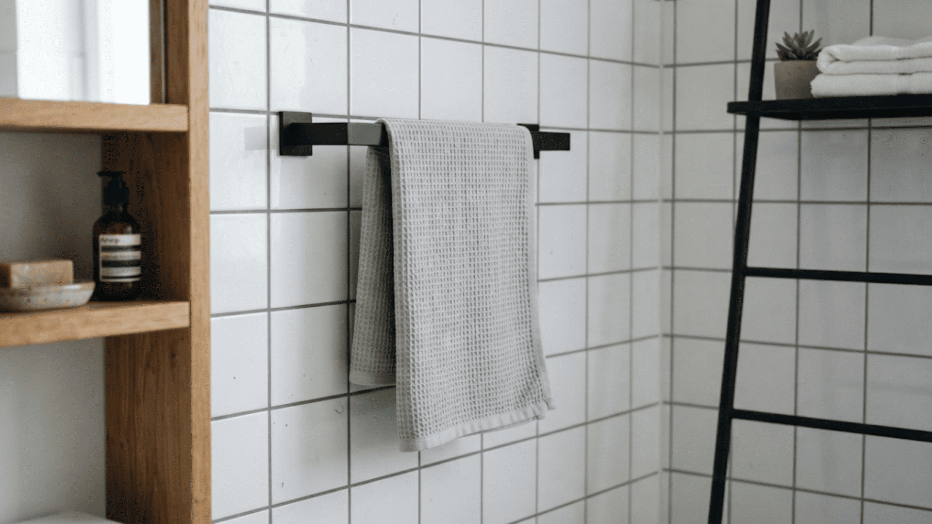 gray waffle mildew smelly towel hanging on black towel bar against white tiled bathroom wall beside wooden shelf