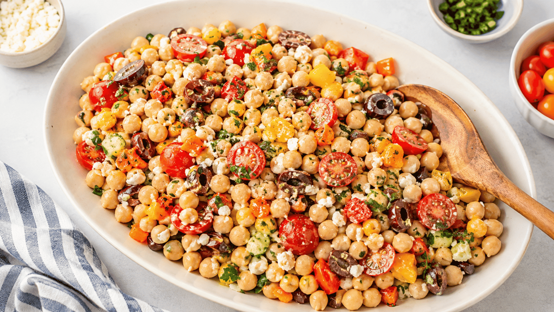 large white platter of chickpea salad with tomatoes olives peppers feta cucumber herbs and a wooden serving spoon beside it