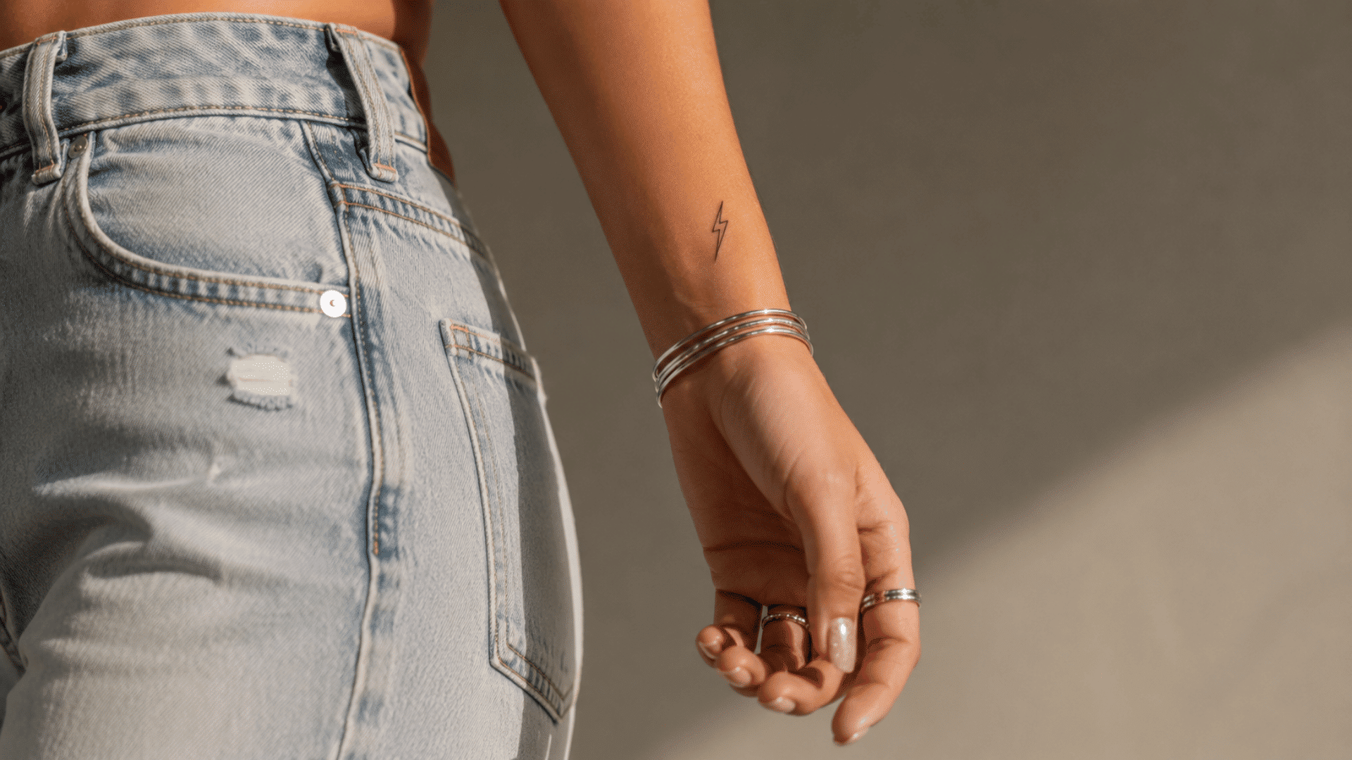 lightning bolt tattoo on the wrist with silver bangles, rings, and light-wash denim jeans.