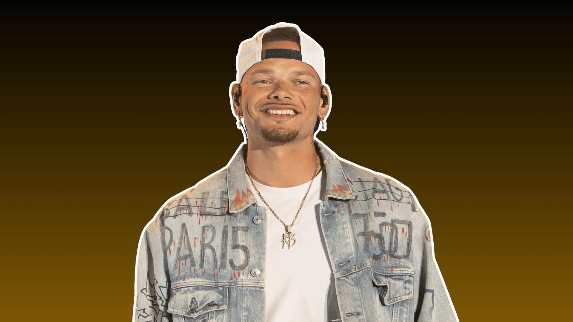 man wearing a backward cap and denim jacket smiling in a portrait against a dark brown background