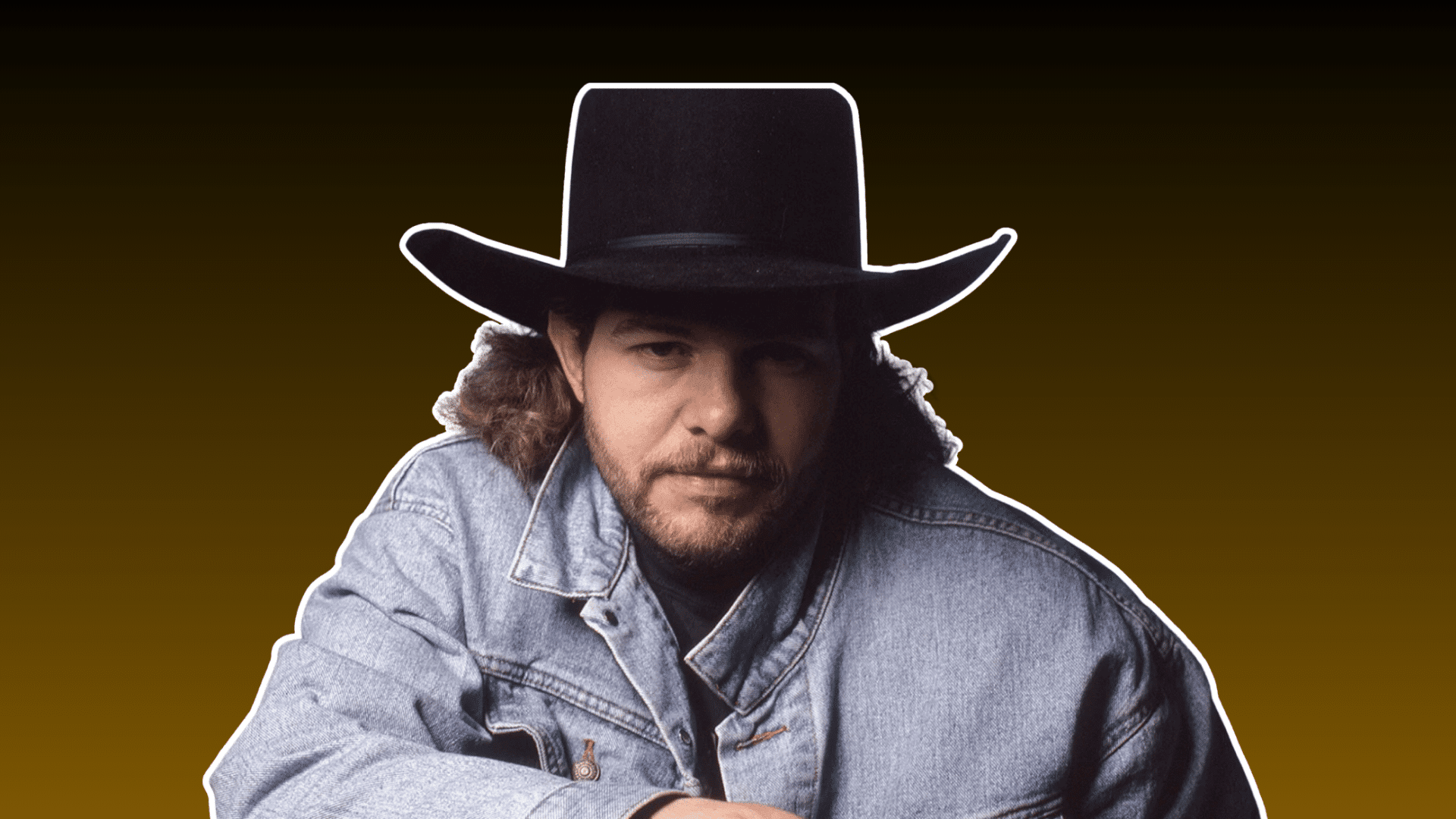 man wearing a black cowboy hat and denim jacket posing against a dark brown background