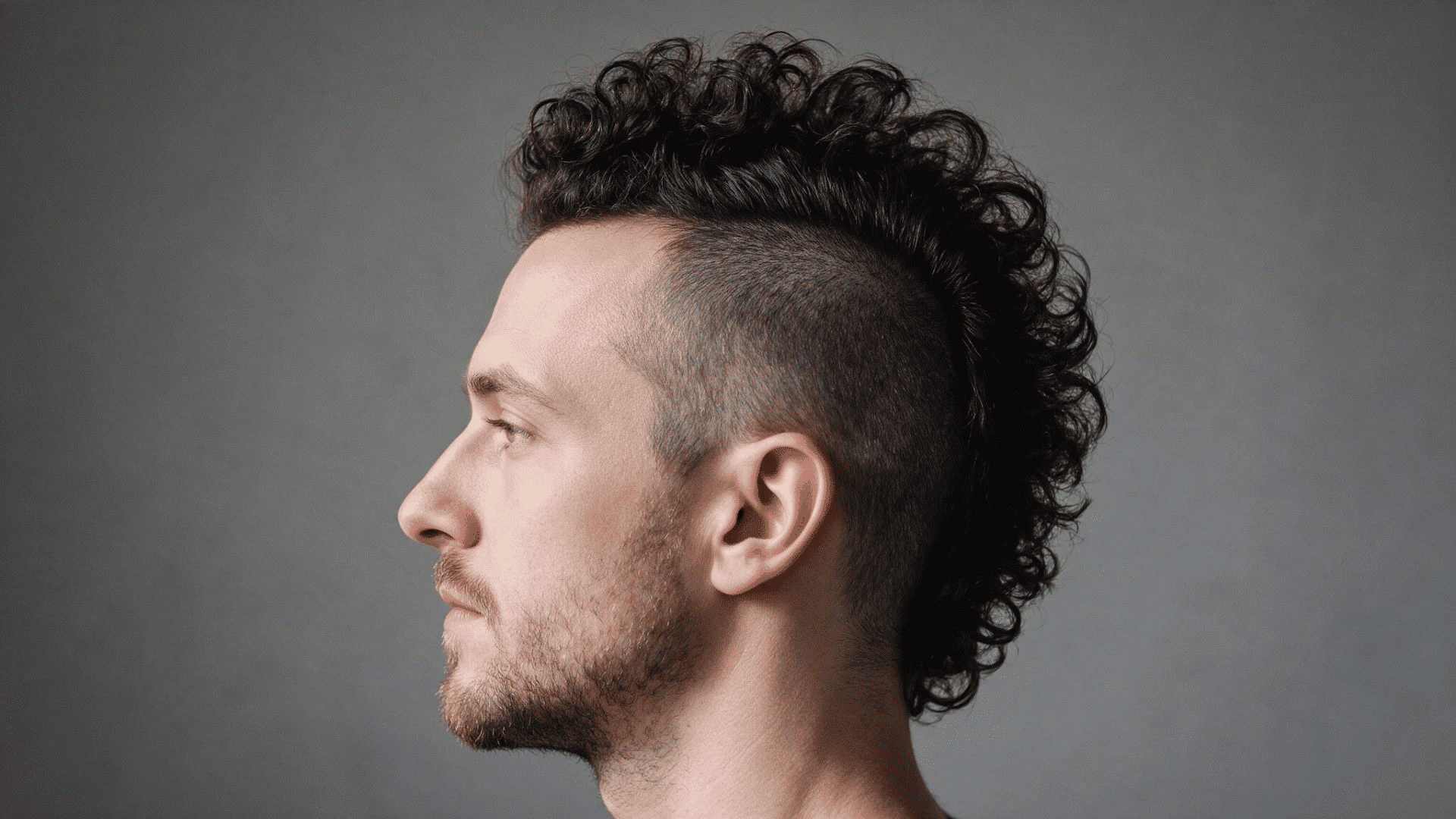 man wearing a deathhawk with a wide strip of dark curly hair through the center and closely shaved hair along both sides