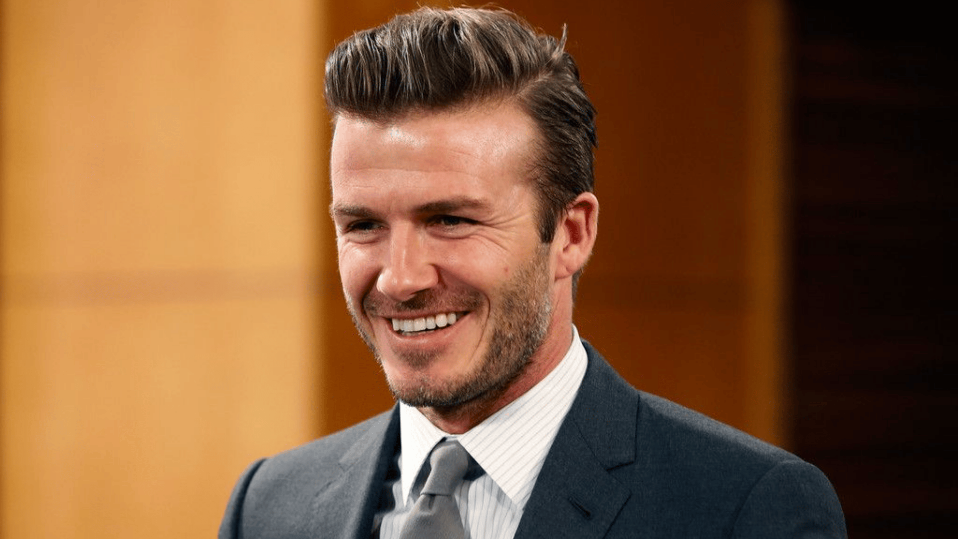 man wearing an ivy league haircut with dark suit striped shirt gray tie and light stubble while smiling at a formal event